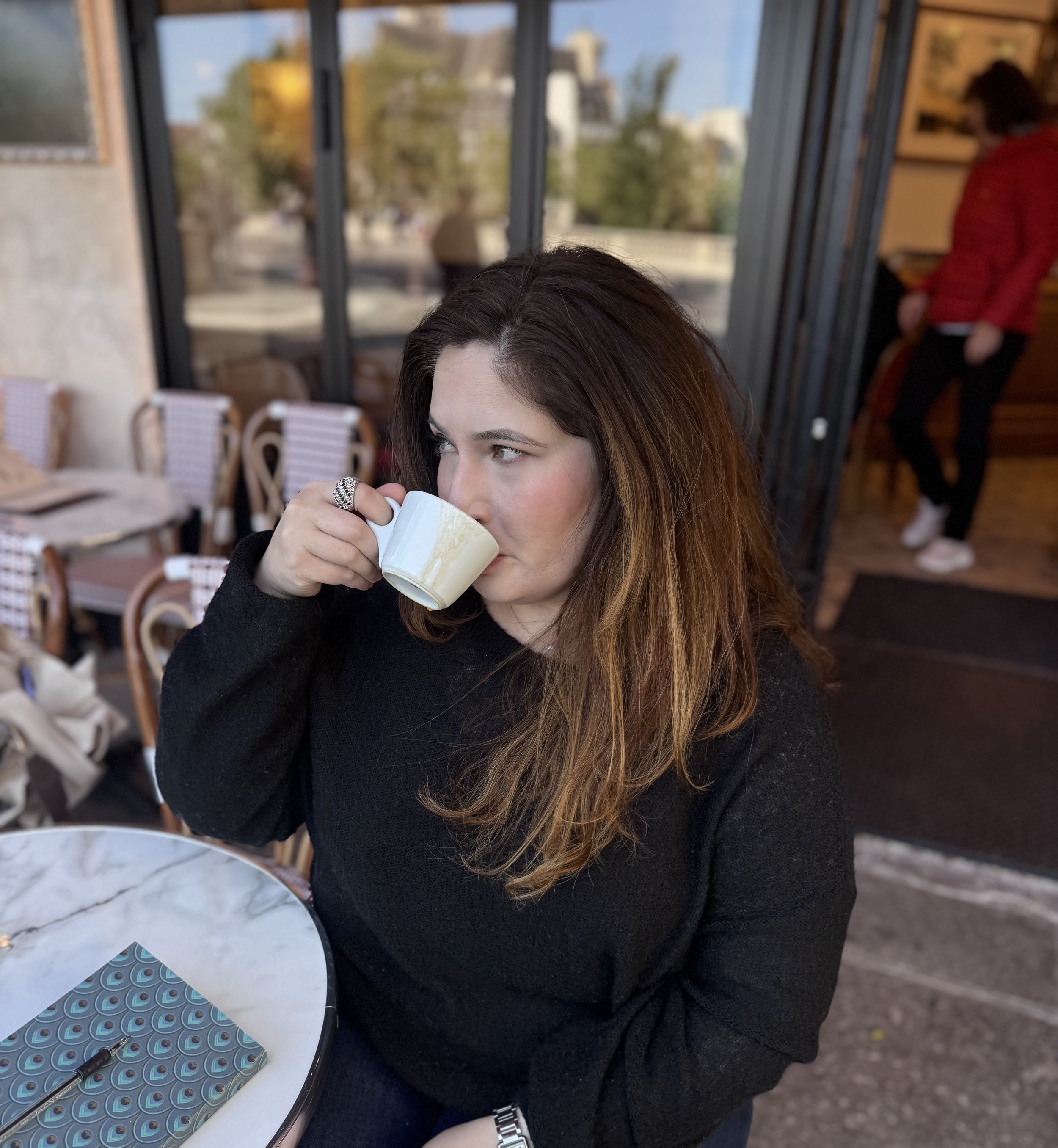 A woman with long, brown hair and a black sweater is sitting at a table, drinking from a white mug. She is wearing a silver watch on her left wrist and a large ring on her right hand. In the background, a person in a red jacket is standing near a wooden wall. The setting appears to be a cozy indoor space with large windows and wooden furniture.