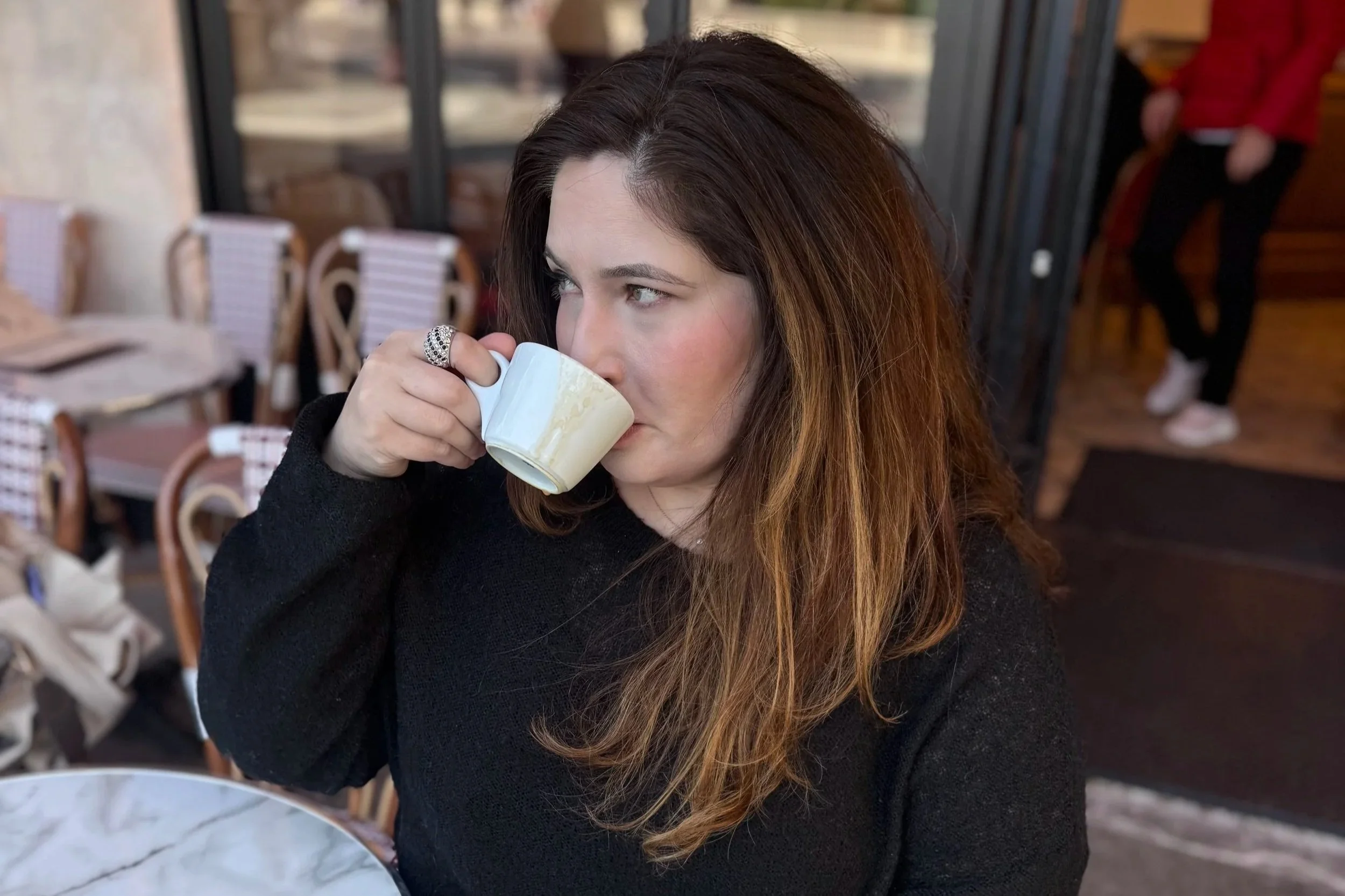A woman with long brown hair drinking from a white cup in a restaurant or cafe.