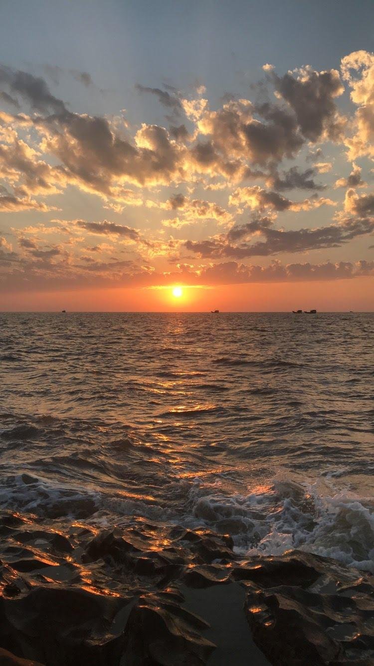 Sunset over ocean with partially cloudy sky and rocky shoreline.