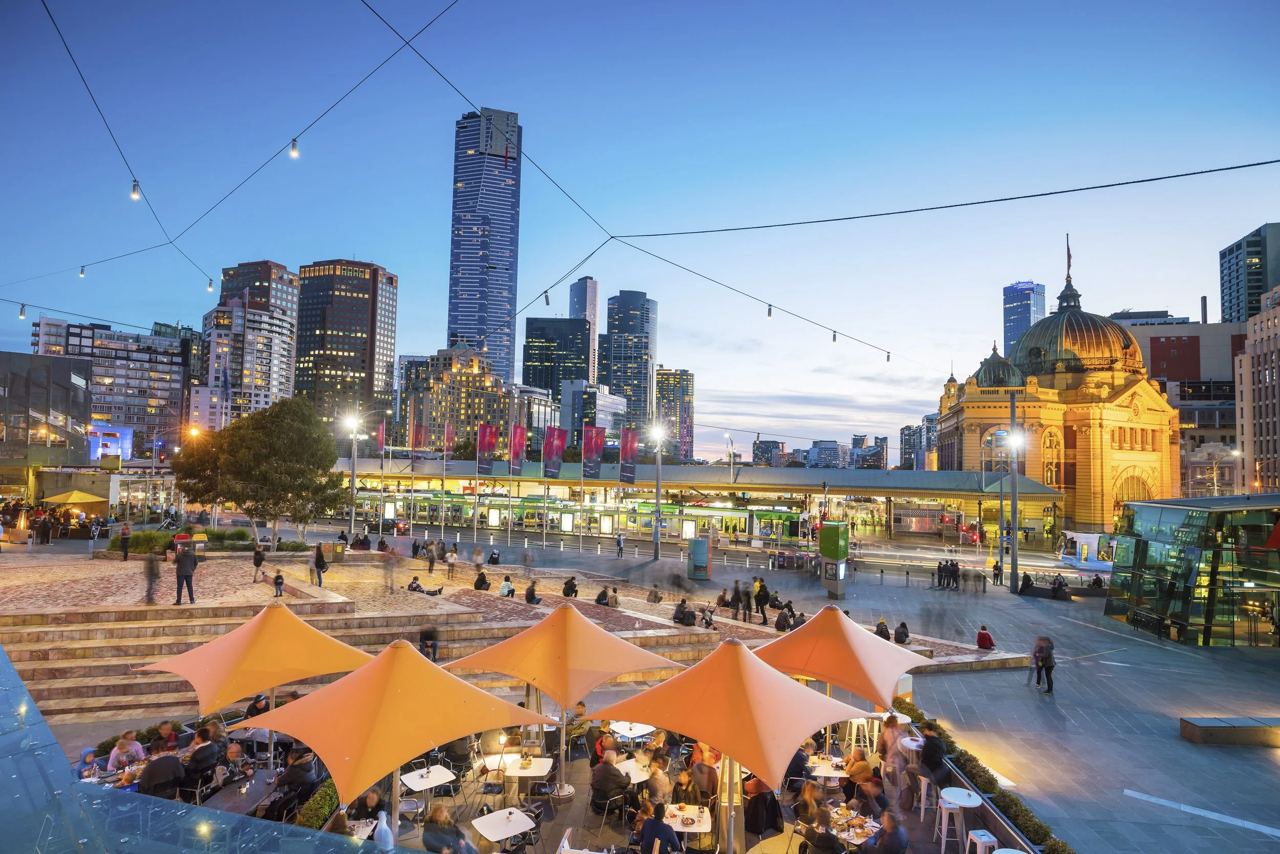 Cityscape of Melbourne, Australia during evening with skyscrapers, historic building, outdoor seating with umbrellas, and people dining.