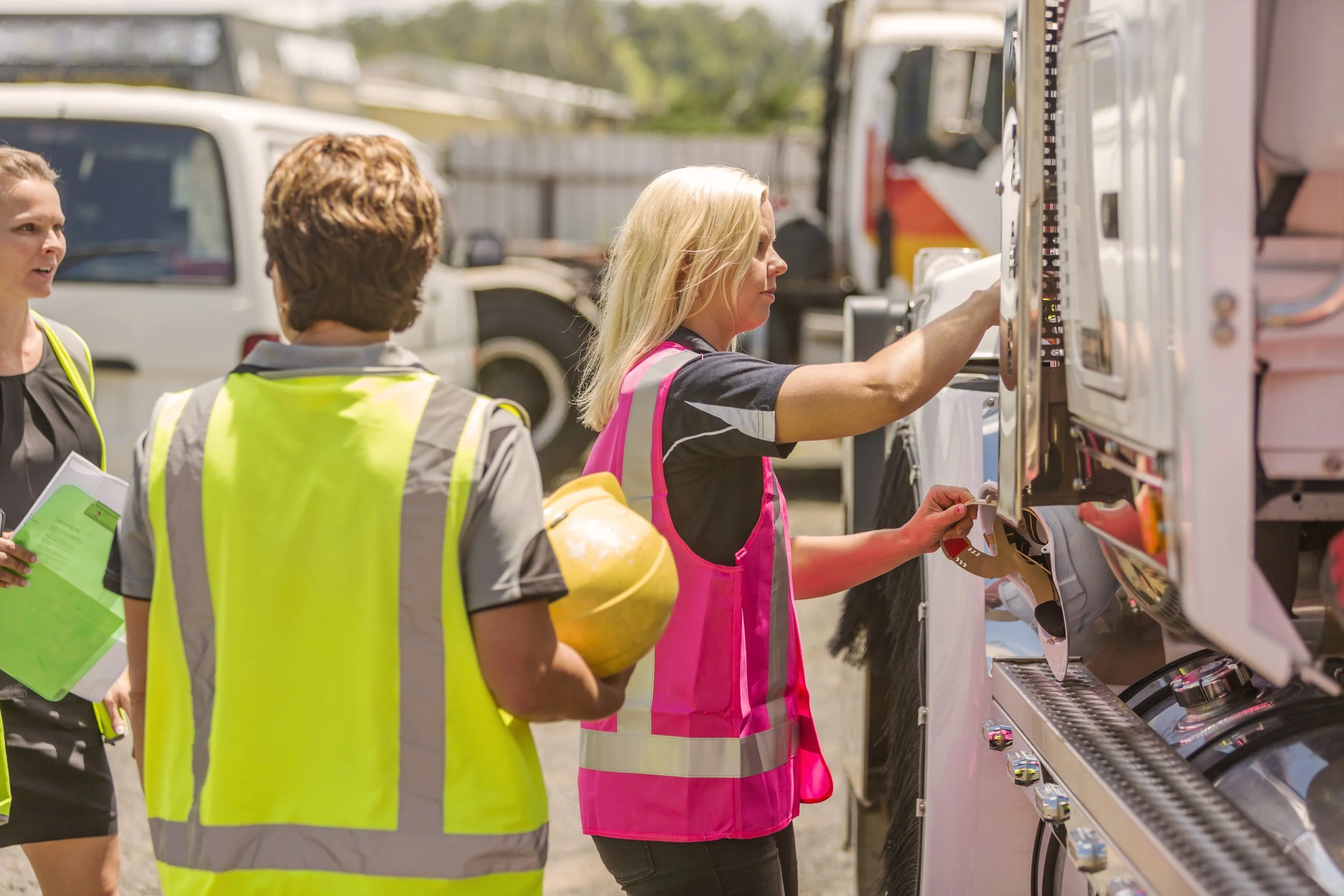 Three women, two in safety vests, are standing outdoors next to fire trucks. One woman is adjusting equipment on the truck, while the other two observe. One of them holds a clipboard, and another has safety helmet. The scene appears to be a safety in