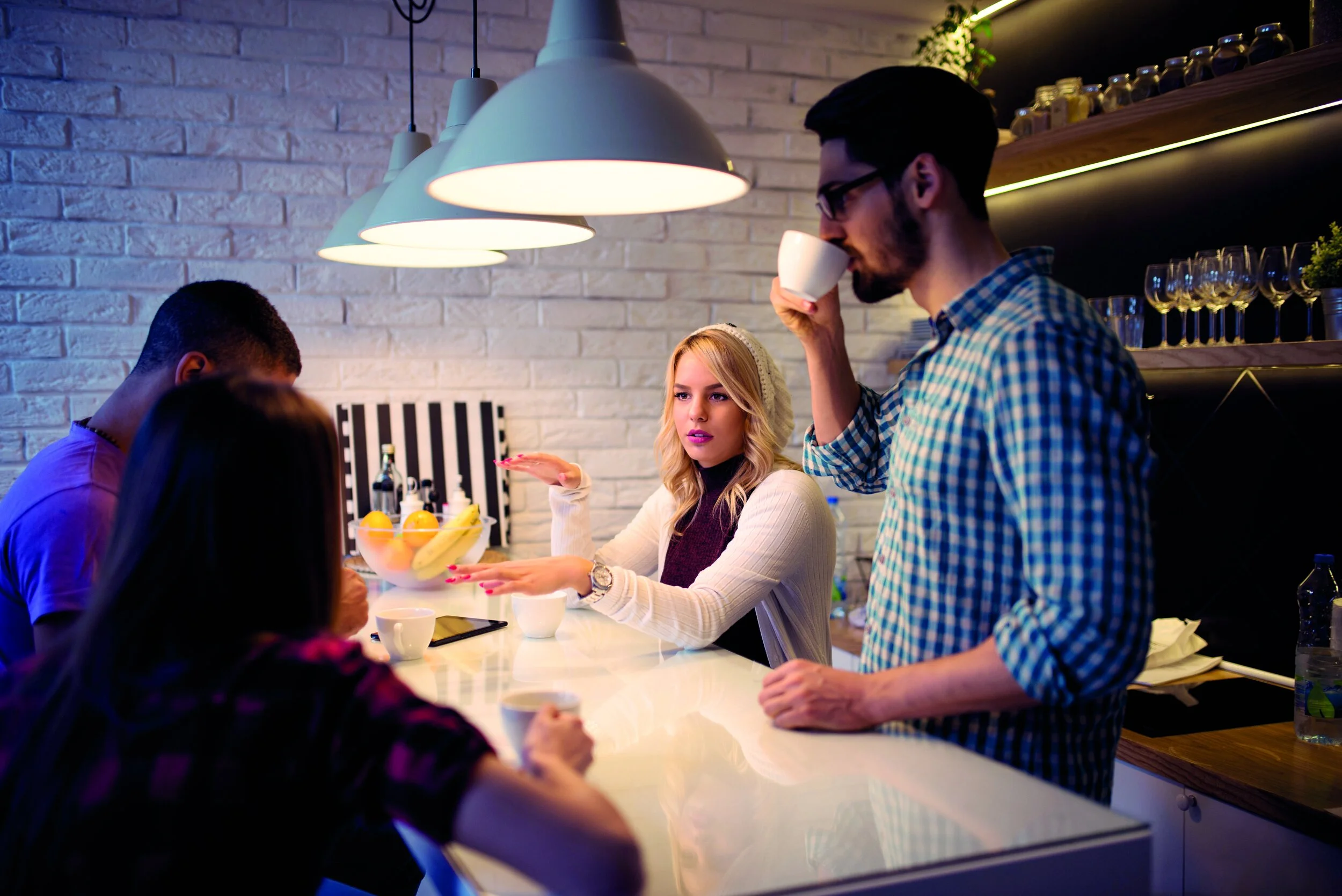 People socializing in a modern kitchen with hanging lamps, a white brick wall, and shelves with glassware and plants. A man drinks from a white cup, a woman gestures while talking, and others are seated at the counter.
