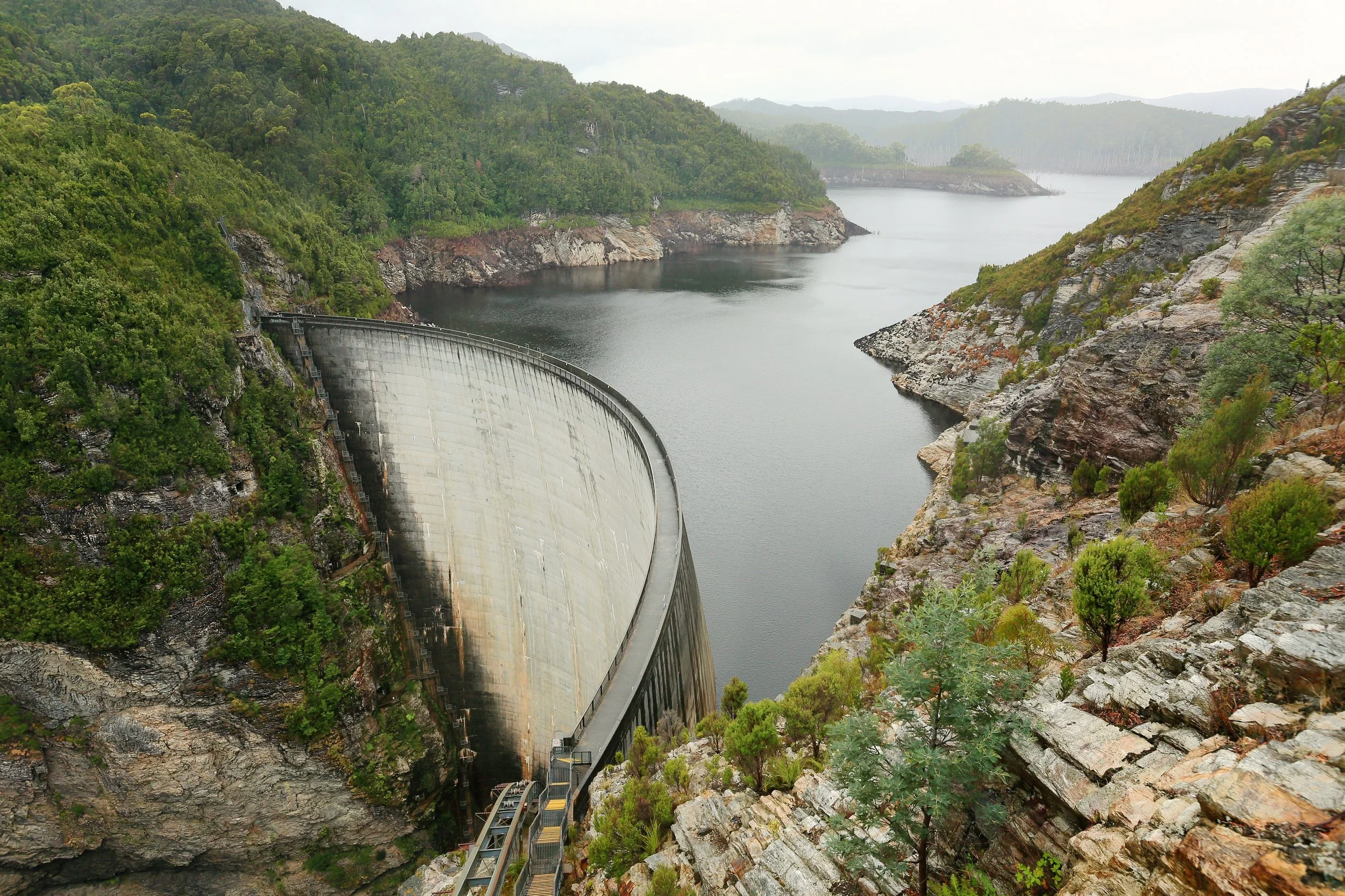 A largeDam with a curved concrete structure spanning between rocky and green hills, with water behind and surrounding landscape of hills and trees.