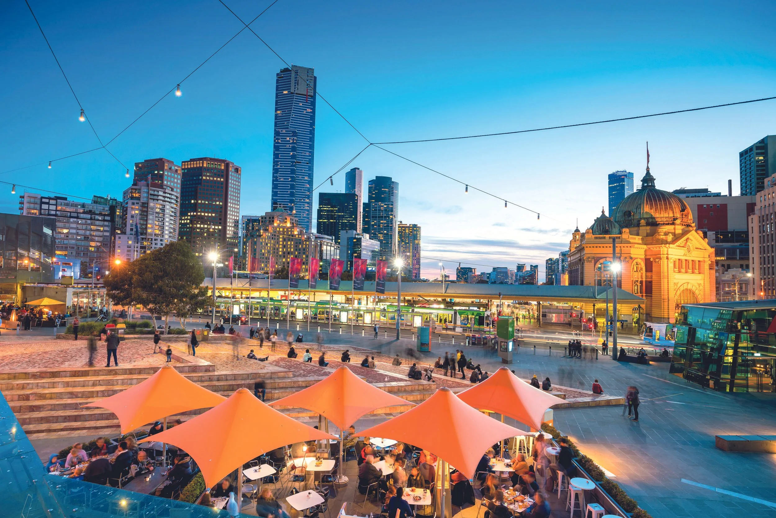 Urban cityscape evening scene with a mix of modern and historic buildings, outdoor dining area with orange umbrellas, people sitting and walking in the plaza, and a train station in the background under a blue sky.
