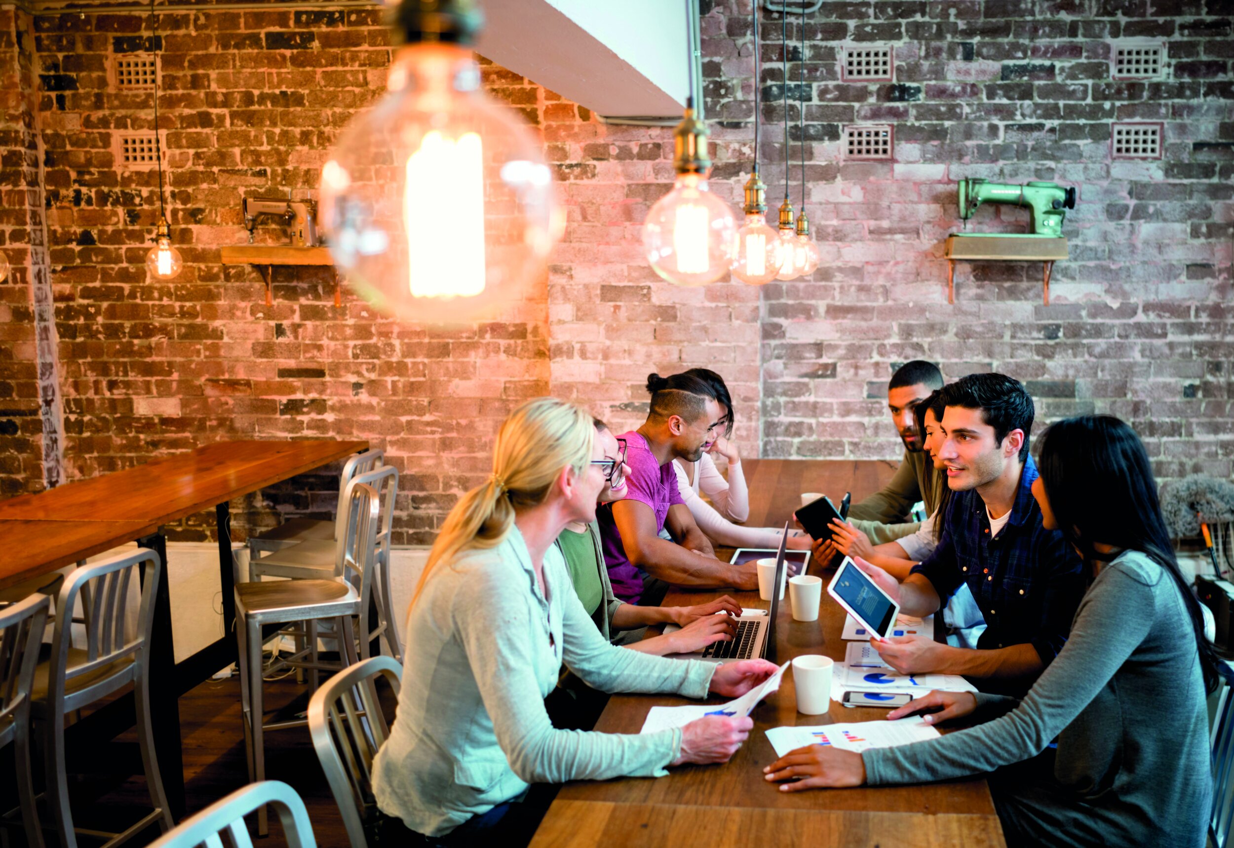 Group of six diverse people sitting at a long wooden table in a modern, brick-walled room, engaged in a discussion with laptops, tablets, papers, and coffee cups. Hanging Edison-style light bulbs overhead illuminate the scene.