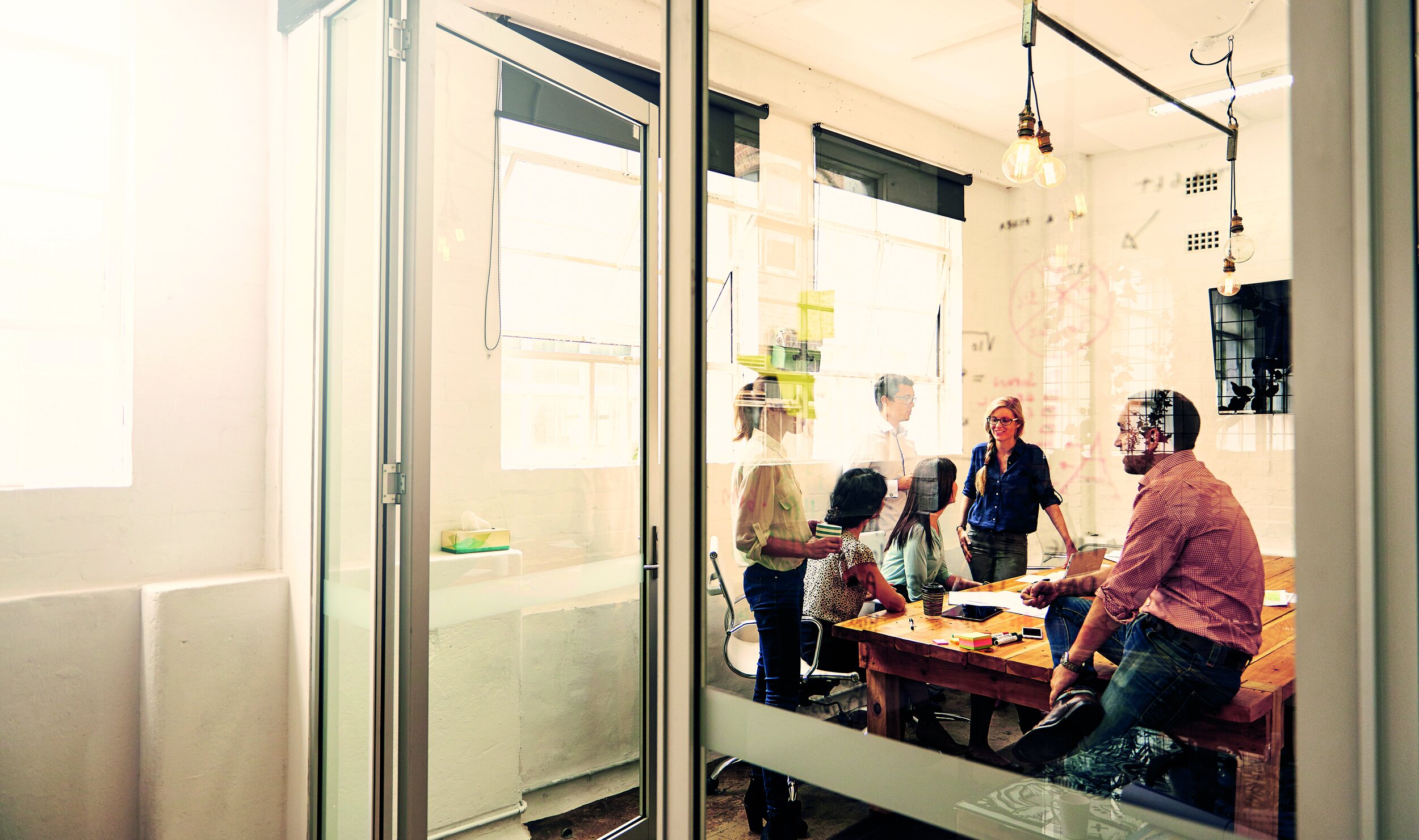 A group of people in a modern office meeting room, seen through glass windows, engaged in a discussion around a wooden table.