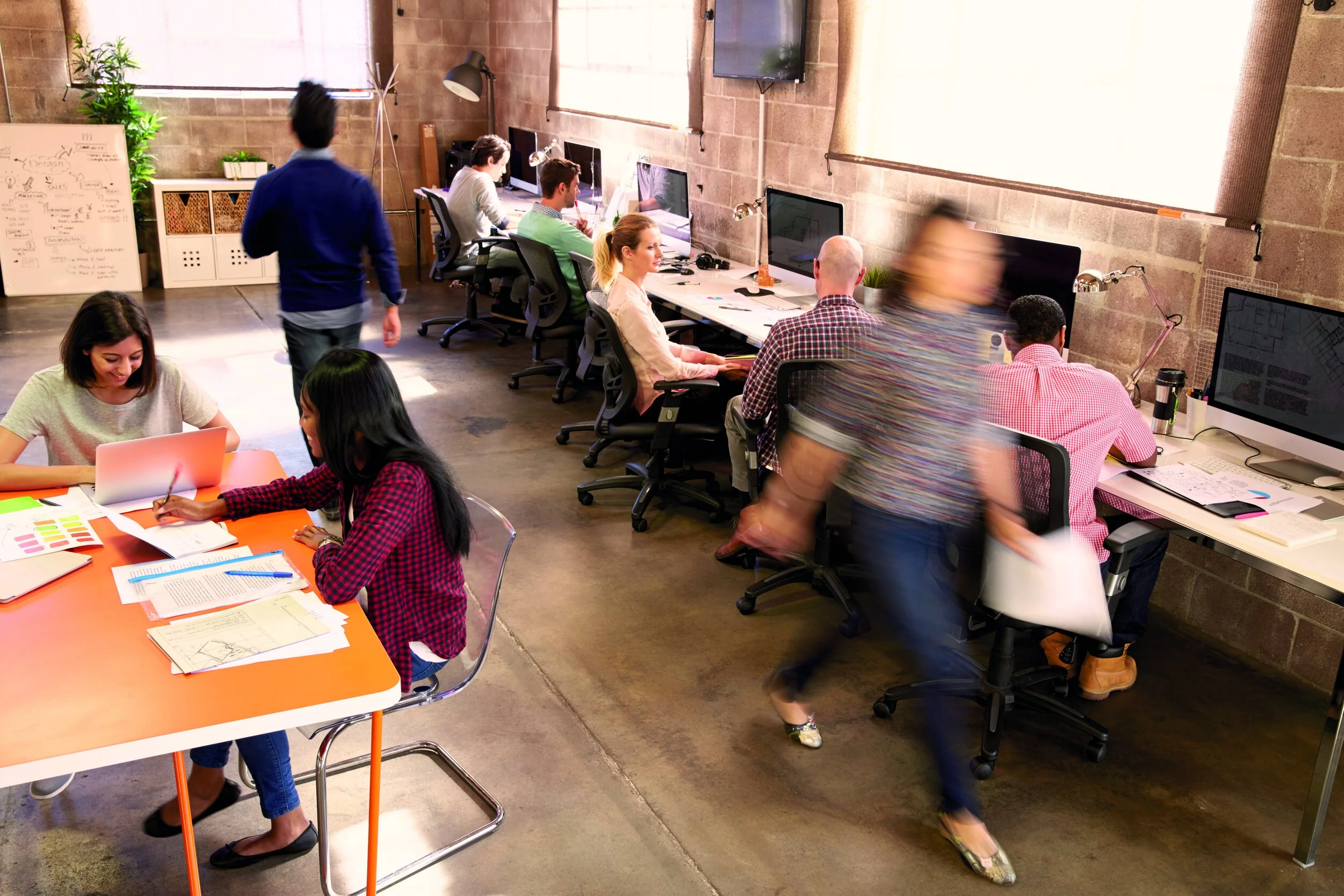 Open office space with multiple employees working at desks with computers, a whiteboard, and a potted plant, with a person walking by and another person blurred in motion.