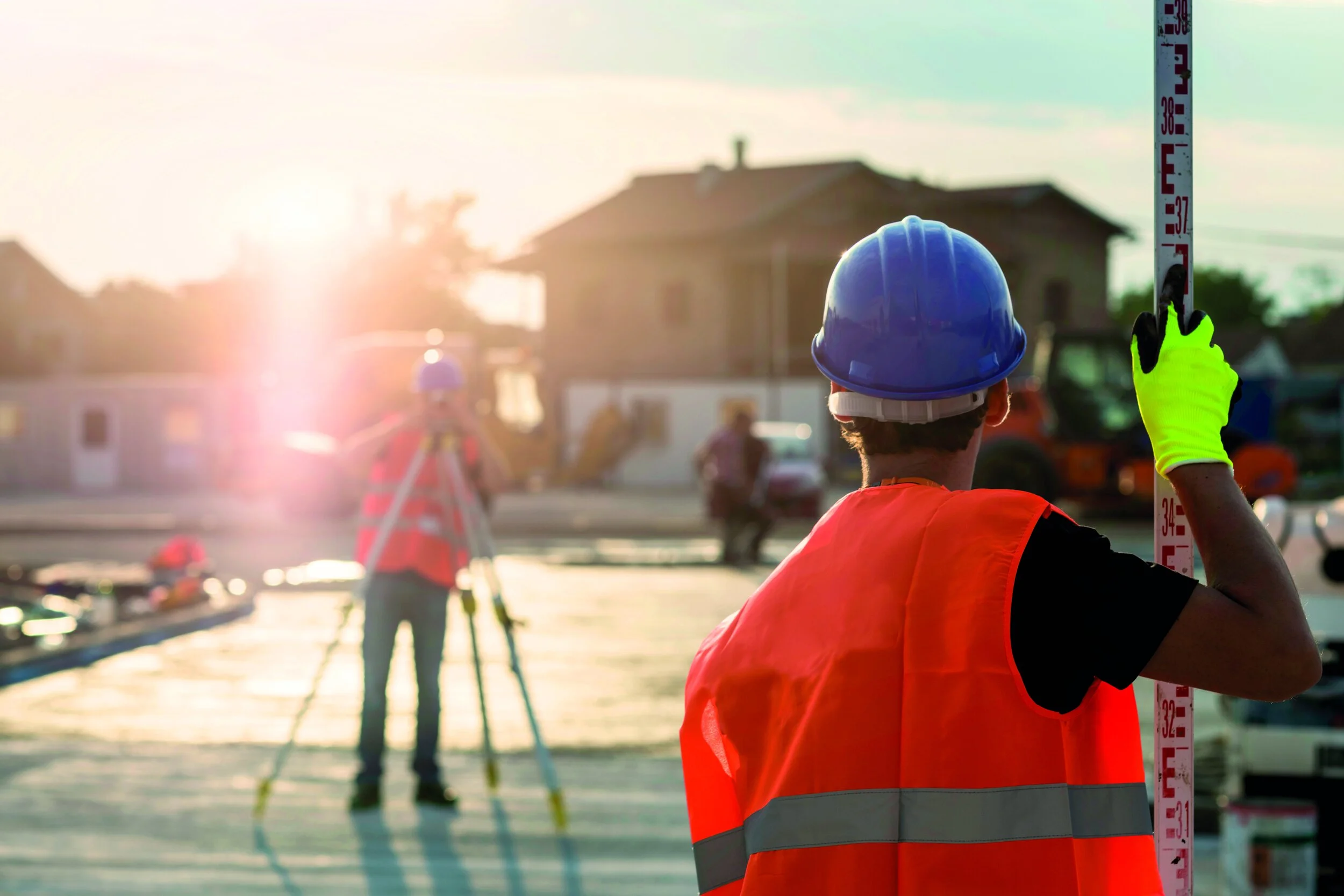 Construction worker in a blue helmet and orange safety vest measuring building height with a level ruler at sunset, with another worker using surveying equipment in the background.