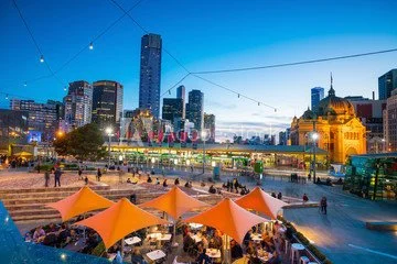 Outdoor event with orange tents, people walking around, in front of a city skyline with tall buildings and a historic-style building, during evening with string lights overhead.