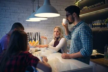 A group of four young adults chatting and drinking coffee or tea at a modern café bar with hanging lights and shelves of glassware.