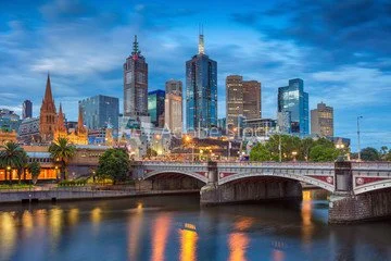 Skyline of downtown city with tall skyscrapers, a bridge over a river, and trees in foreground, at dusk.