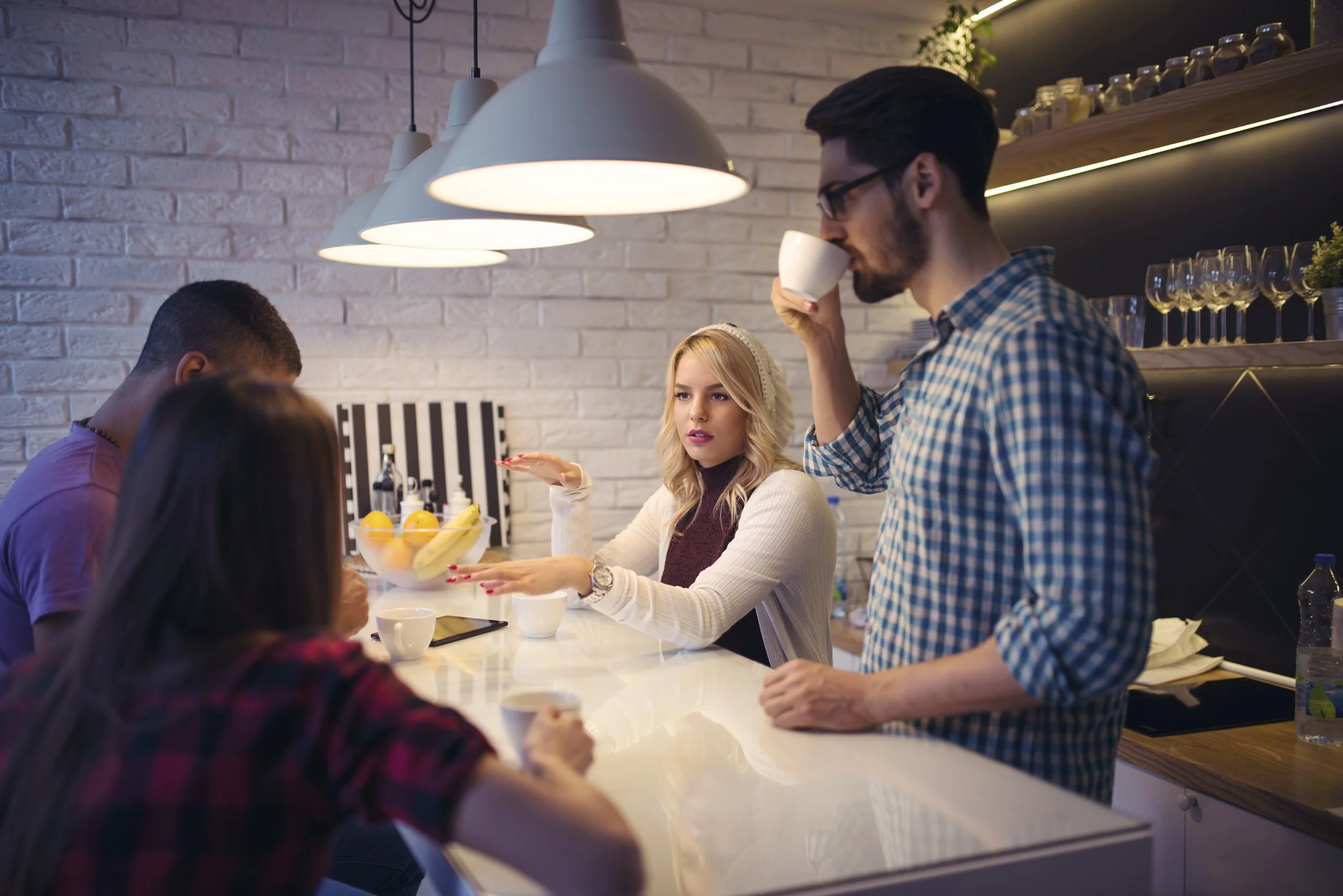 Four people gathered around a kitchen counter in a modern kitchen, with two hanging lamps overhead and a white brick wall in the background. One woman with blonde hair and a white sweater is gesturing with her hands, another woman with dark hair in a