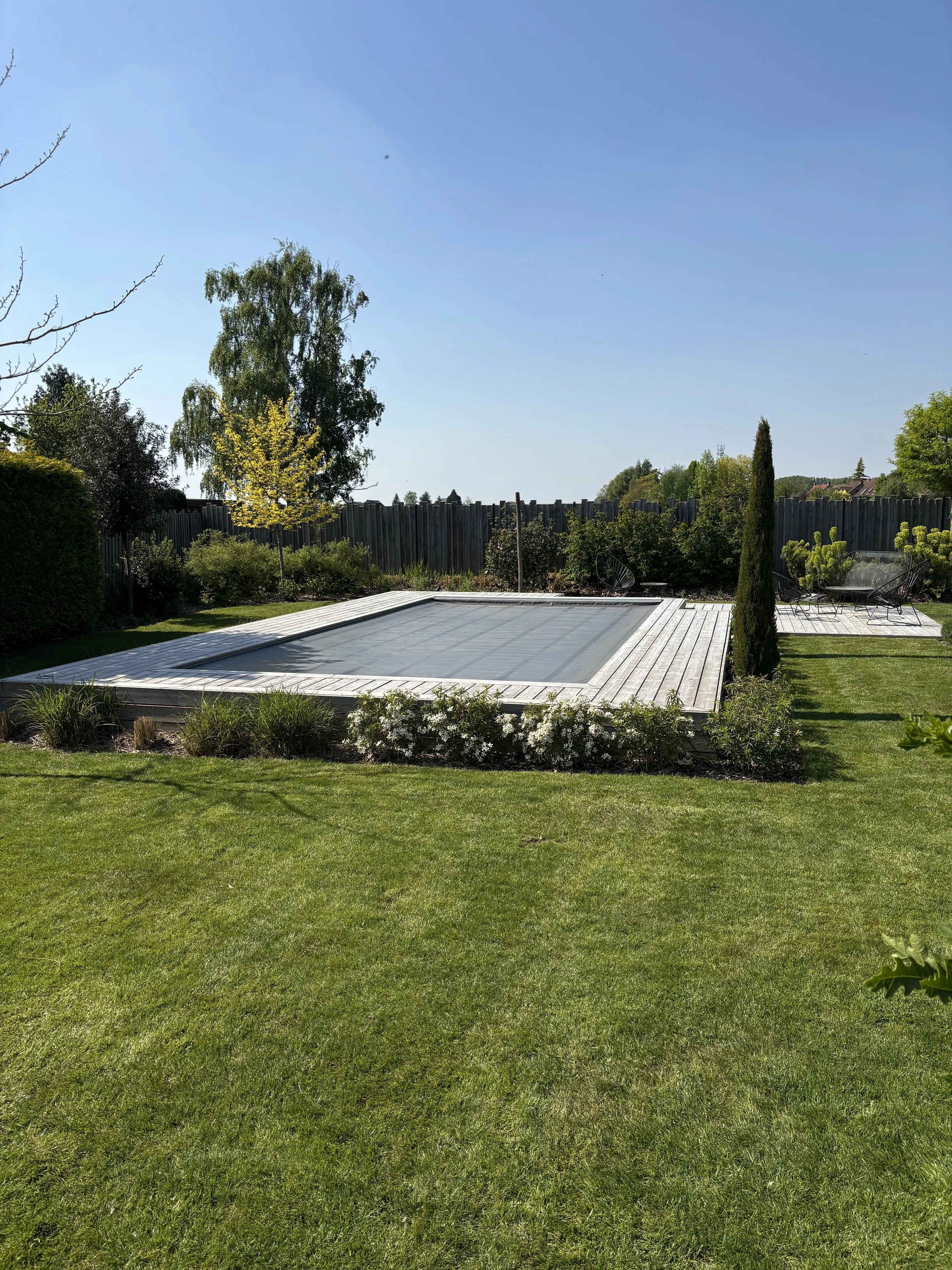 Piscine couverte dans un jardin ensoleillé, entourée de pelouse et de végétation, avec un ciel clair et bleu.
