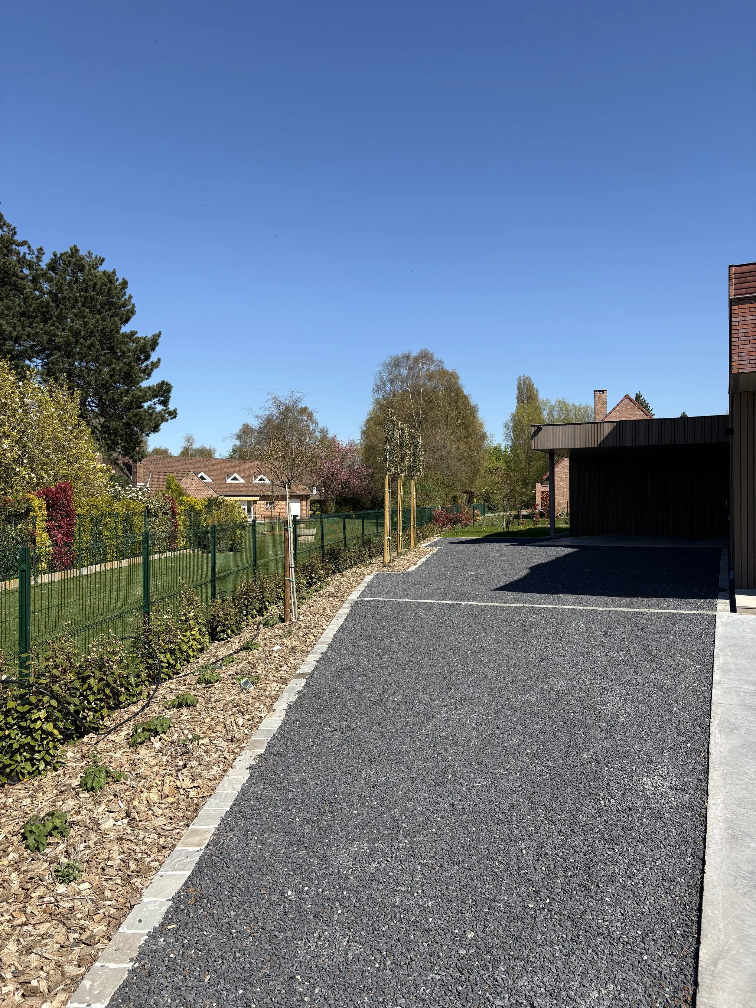 Chemin pavé de gravier noir bordé de petites plantes et d'un trottoir en pierre, avec un jardin clos de clôture verte, des arbres et des maisons résidentielles en arrière-plan sous un ciel bleu clair.
