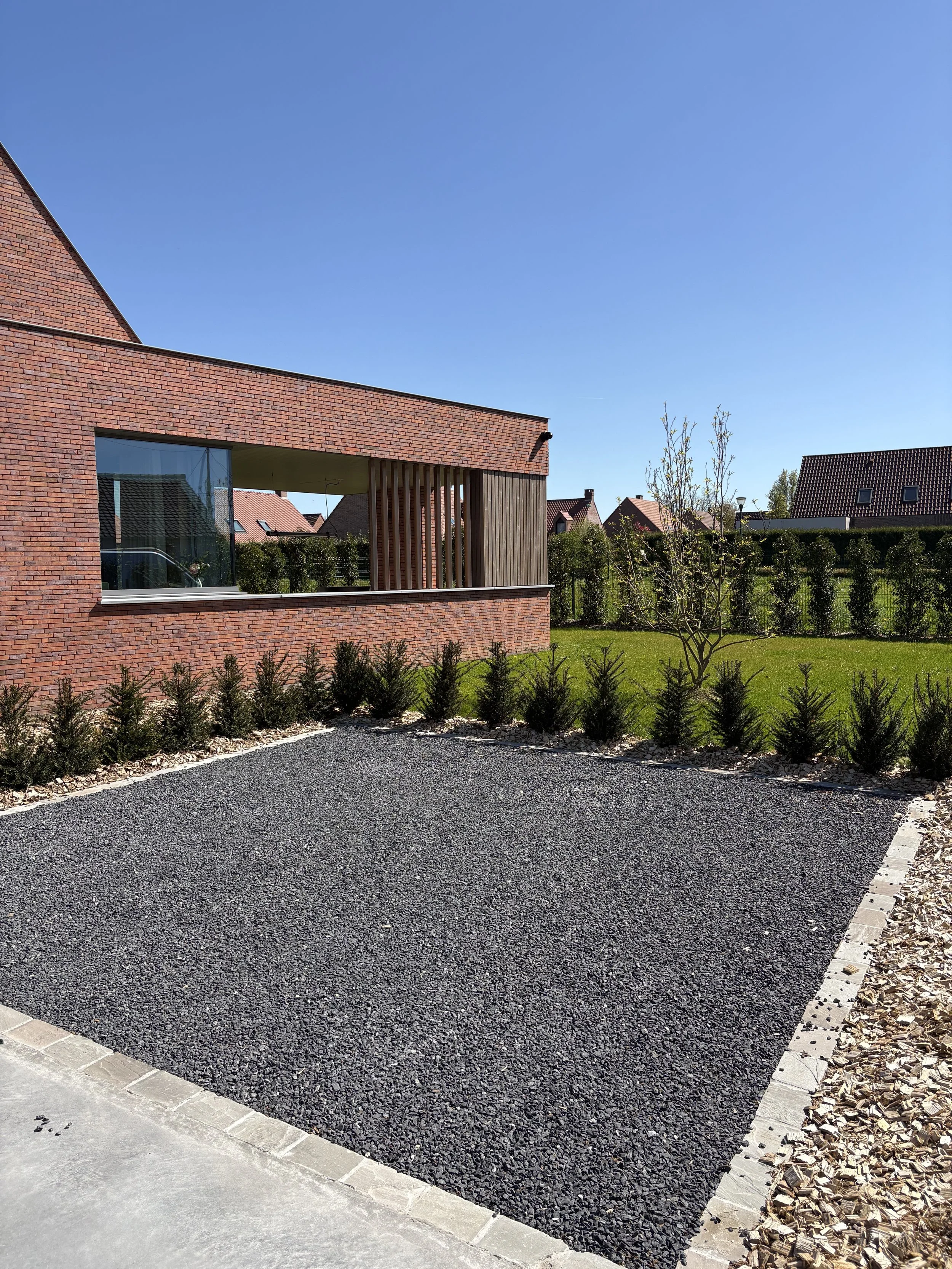 Un parking en gravier noir avec une bordure en pavés, un bâtiment en briques rouges avec une fenêtre en verre, un jardin avec des jeunes arbres et des buissons, sous un ciel clair et ensoleillé.