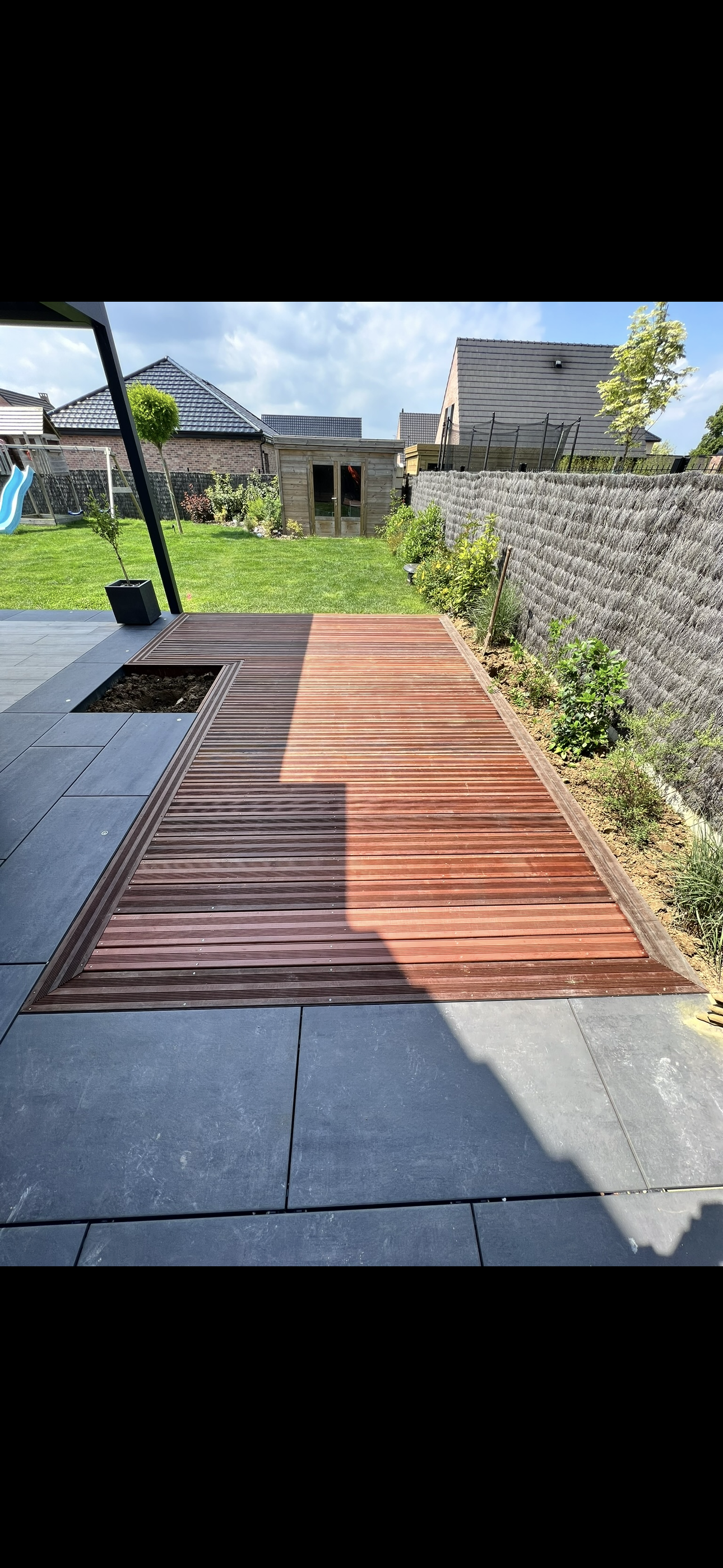 Un jardin avec une terrasse en pavés gris, un solarium en bois rouge et un espace vert avec pelouse, buissons et un petit abri en bois. Foyer de soleil en plastique bleu sur la pelouse.
