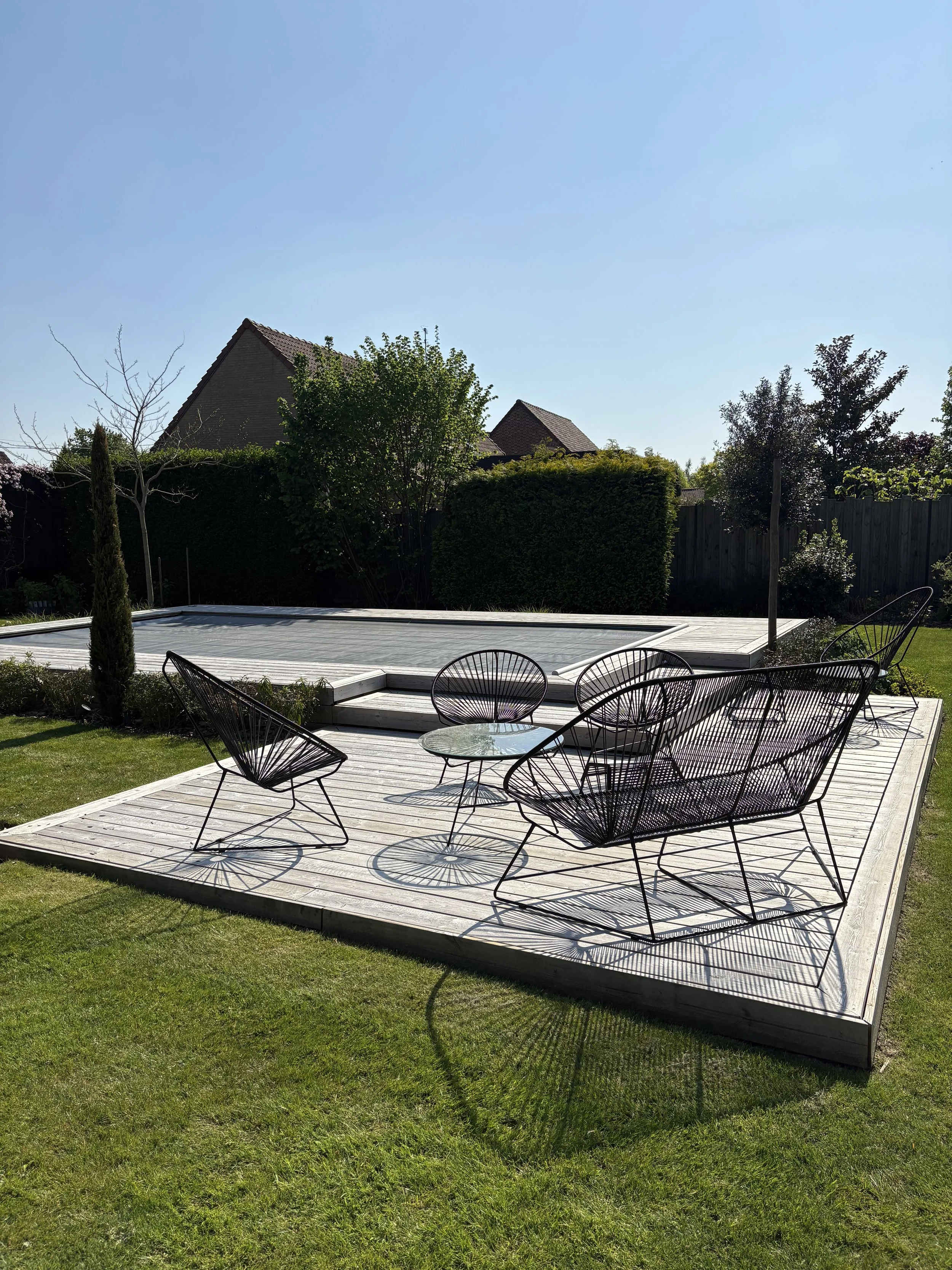 Jardin avec une terrasse en bois, meubles en métal noir, piscine couverte, herbe verte, arbres et clôture en bois sous un ciel clair.