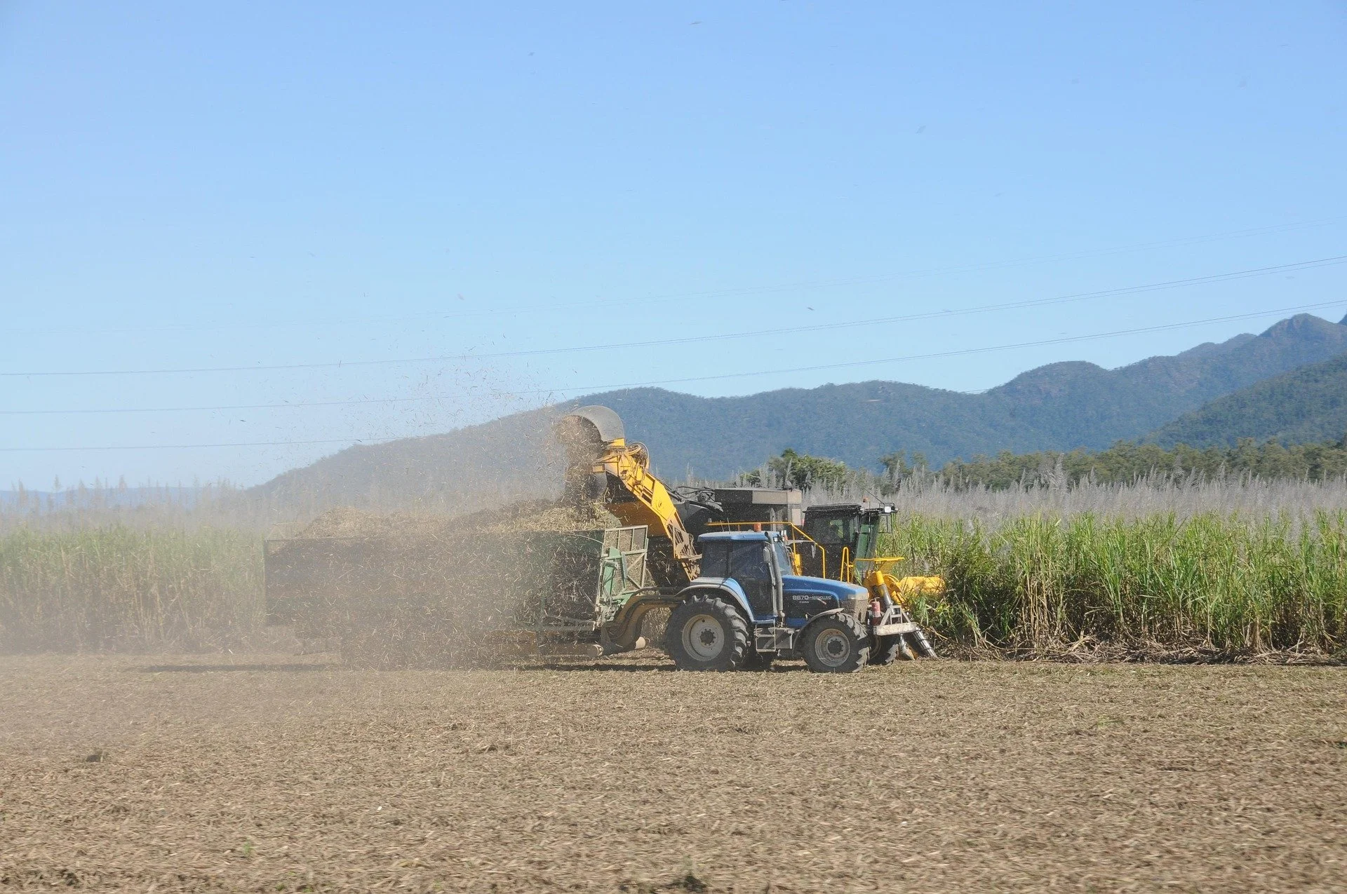 A tractor and harvester working in a farm field, harvesting crops under a clear blue sky, with mountains in the background.