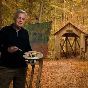 A man standing outdoors in autumn with a palette of paints, beside a large painting on an easel, in front of a wooden gate, surrounded by fall trees.