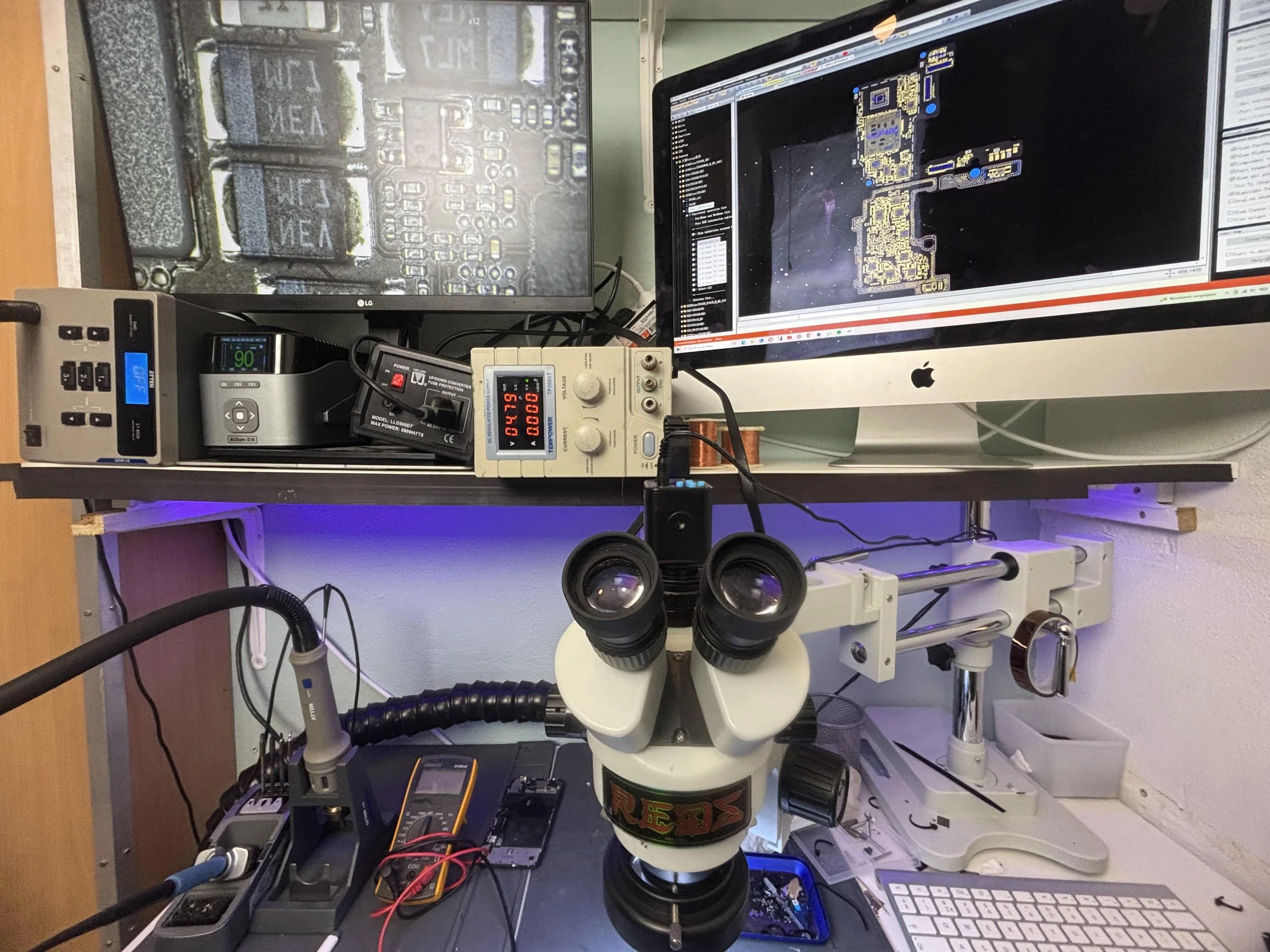 A laboratory workbench with a microscope, electronic testing equipment, a computer monitor displaying circuit design, and electronic components, indicating a workspace for electronics or microscopy work.