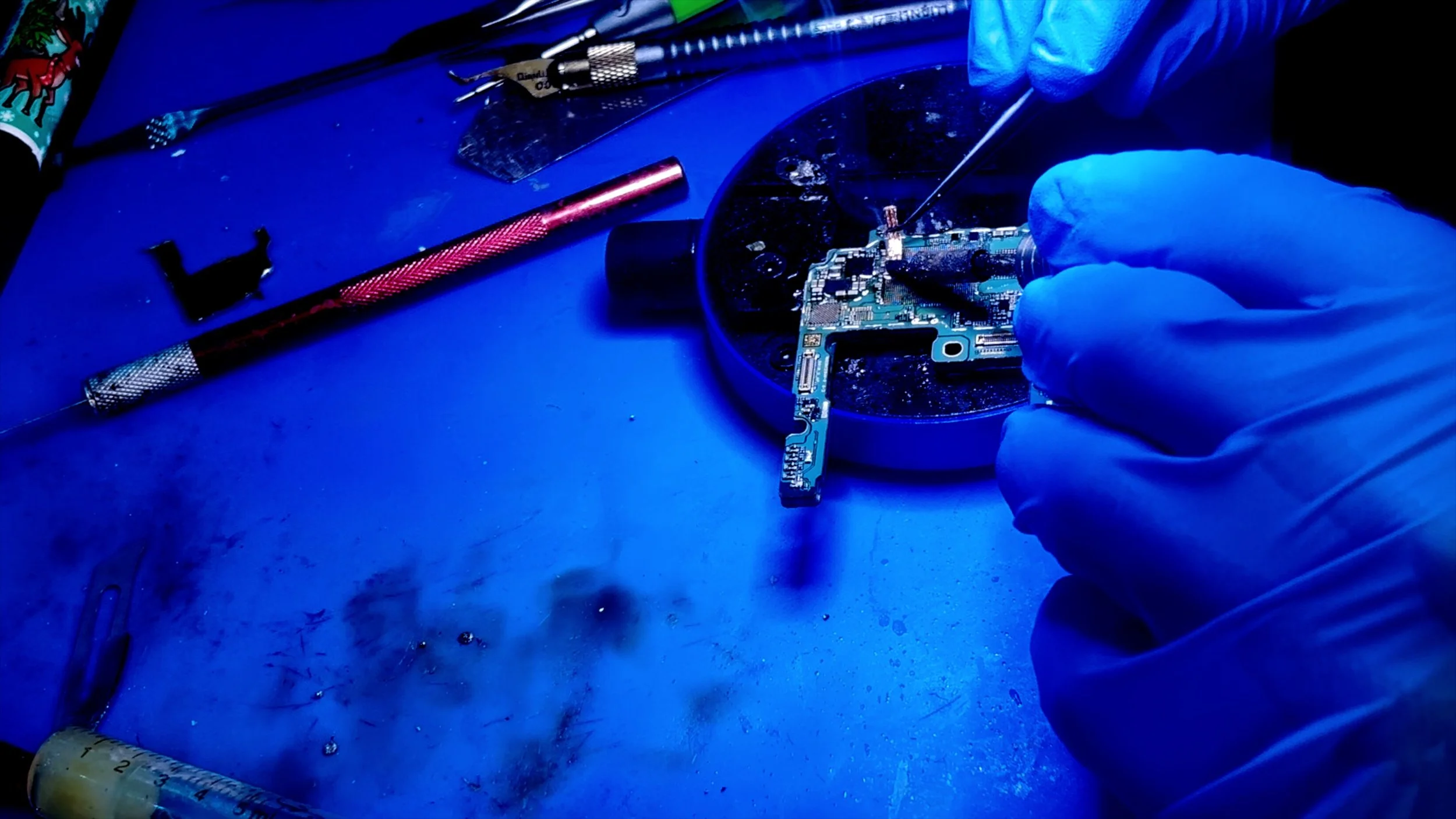 Close-up of a person wearing blue gloves repairing or assembling a circuit board using a precision screwdriver, with tools spread out on a blue work surface.