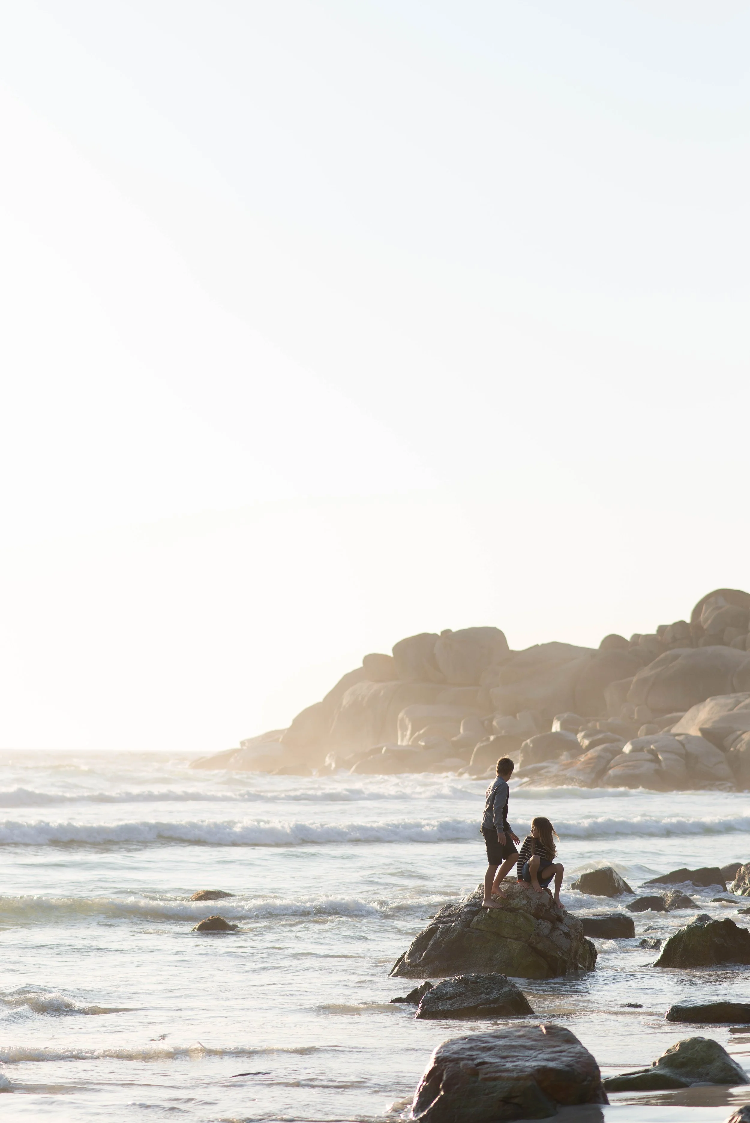 Two children playing on rocks at the beach with ocean waves and rocks in the background, during sunset or sunrise.