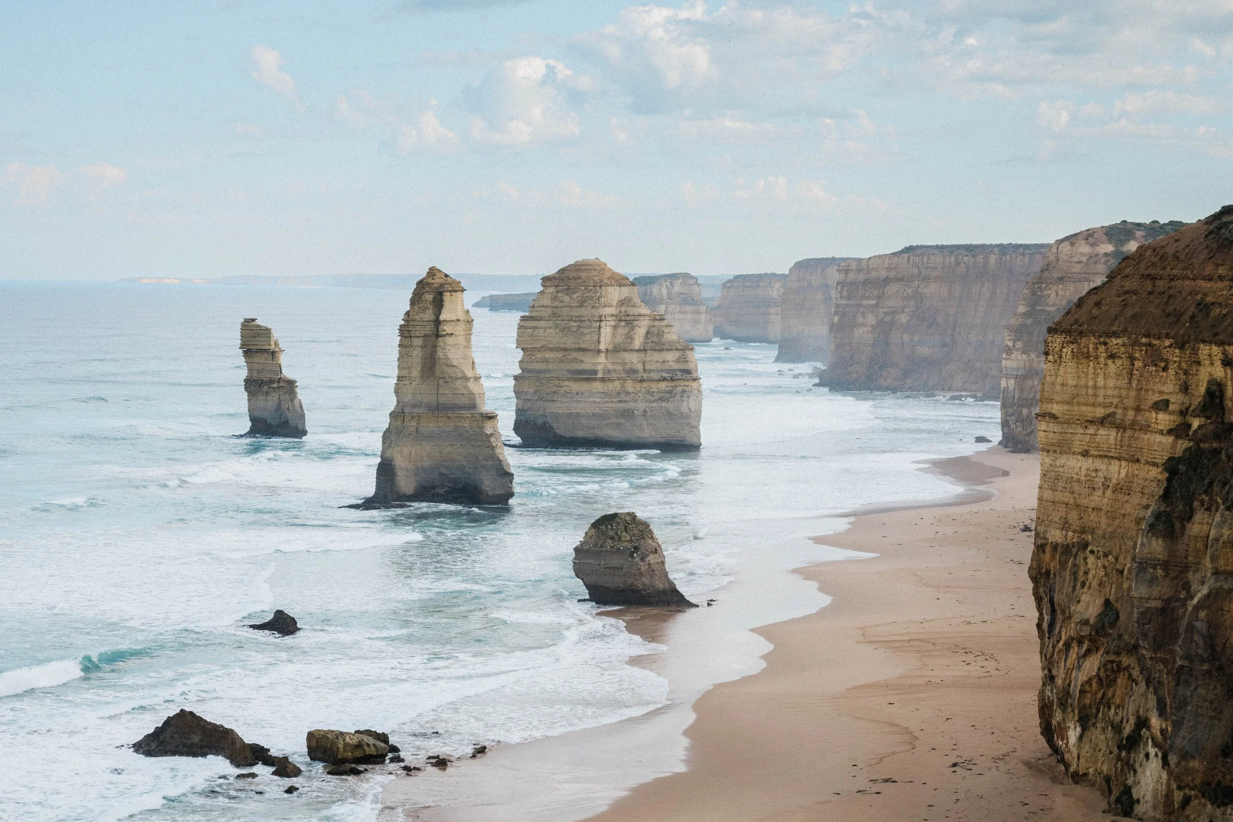 Scenic view of the Twelve Apostles limestone stacks along the Great Ocean Road, with sandy beach, ocean waves, and cliffs under a partly cloudy sky.