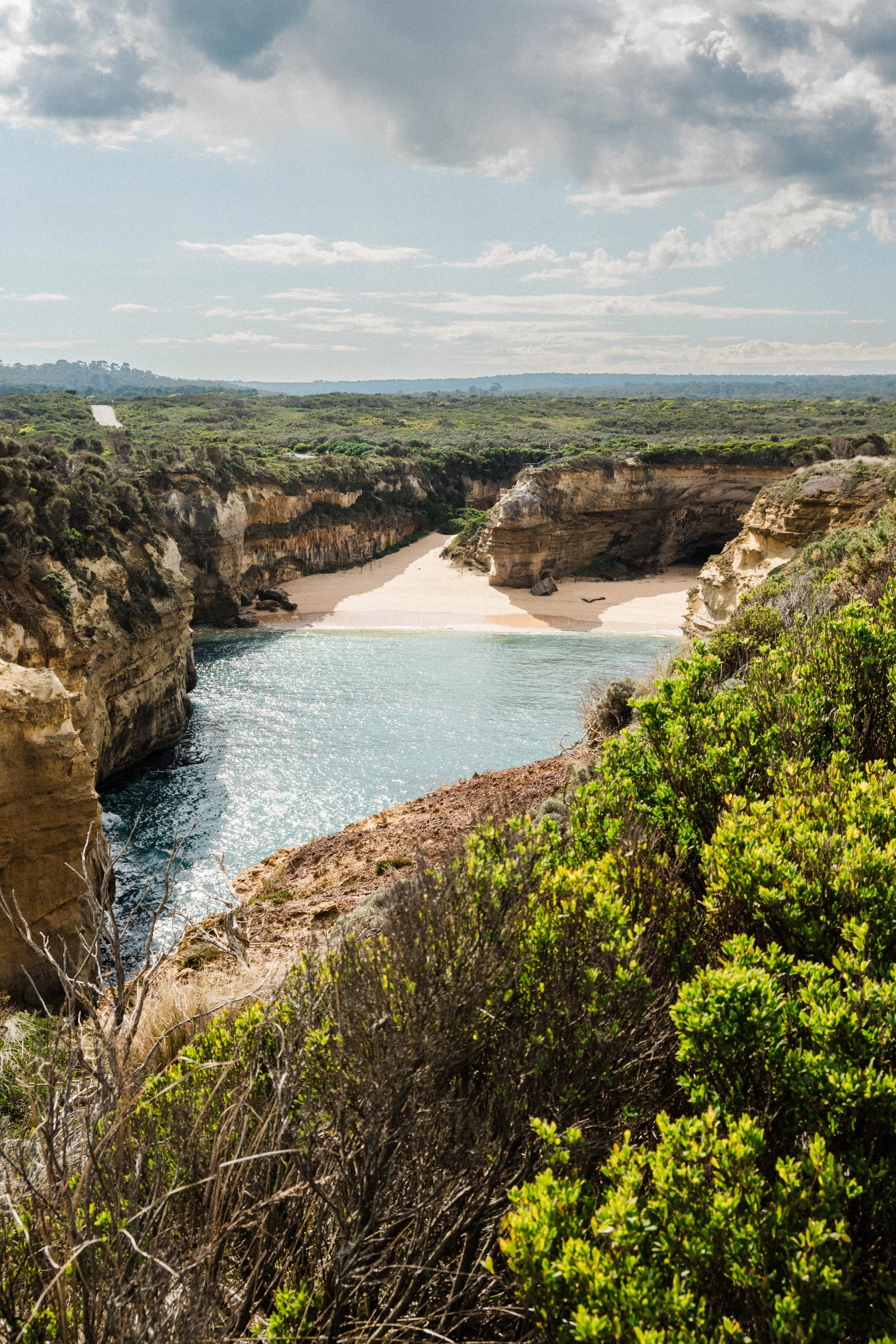 Scenic view of a river flowing between rocky cliffs with sandy shores, surrounded by green shrubbery under a partly cloudy sky.