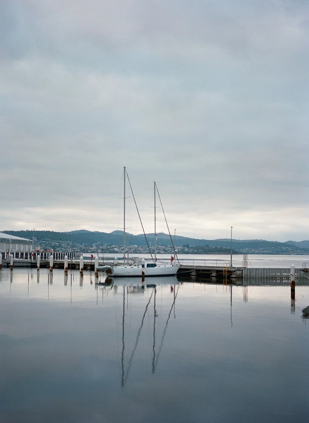 A peaceful harbor scene with sailboats docked at a pier, calm water reflecting the boats and overcast sky, with hills and buildings in the background.