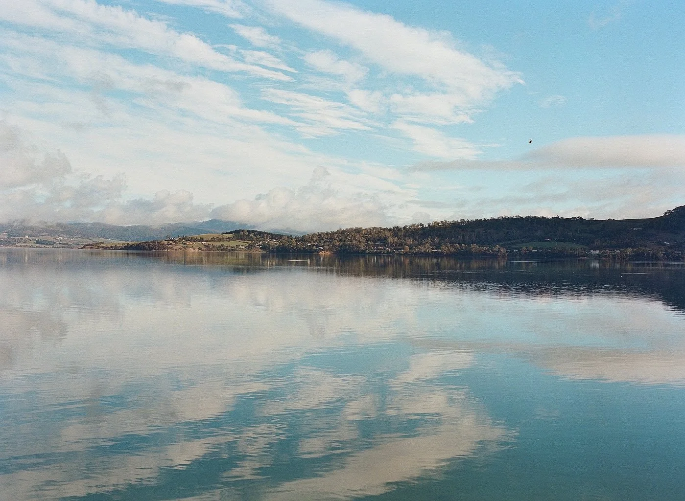 Scenic view of a calm lake with reflections of the sky and distant hills, some clouds, and a bird flying in the sky.