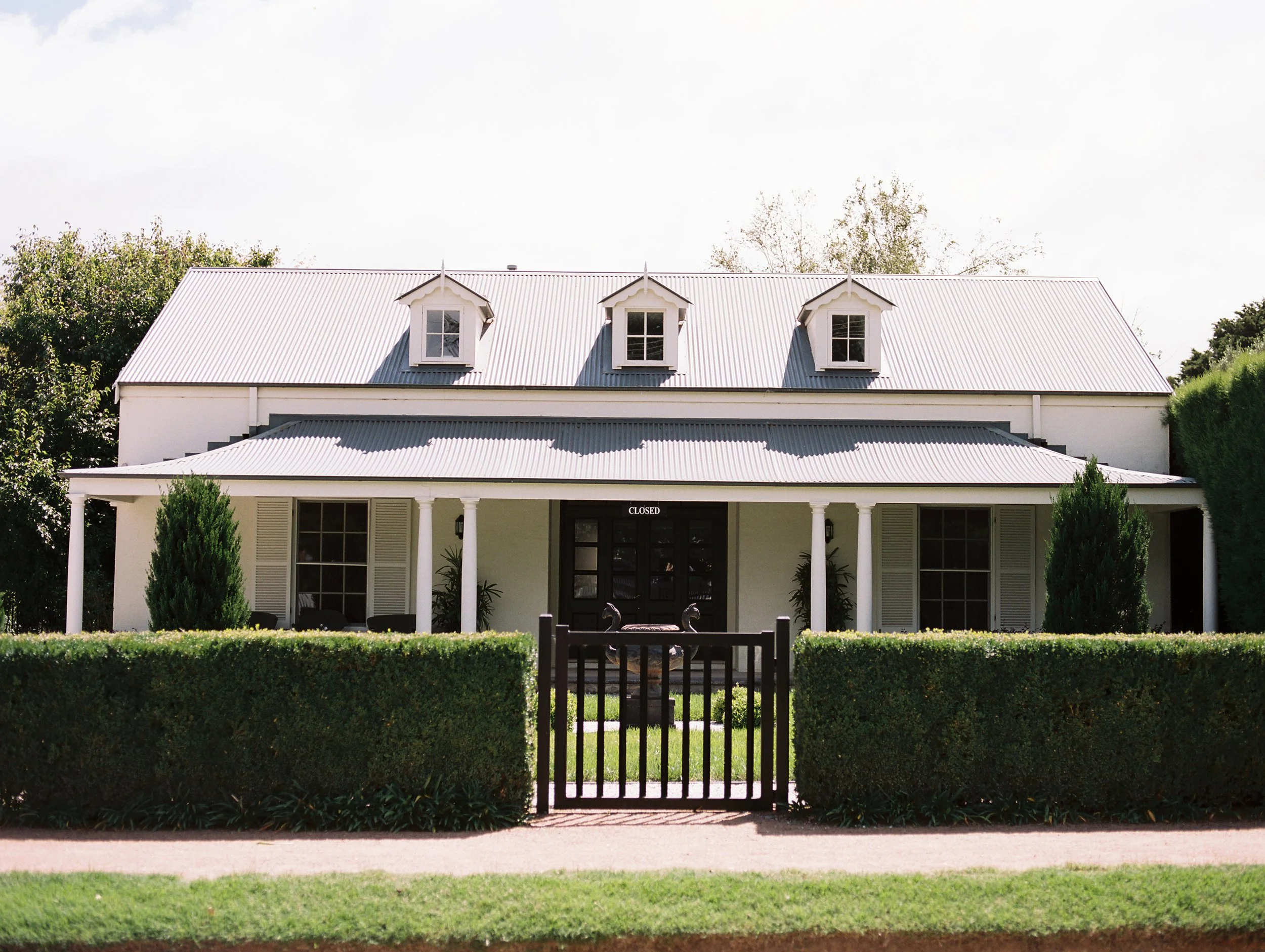 White house with three dormer windows, black front door with a "CLOSED" sign, surrounded by trimmed hedges and trees, with a decorative black metal gate at the front.