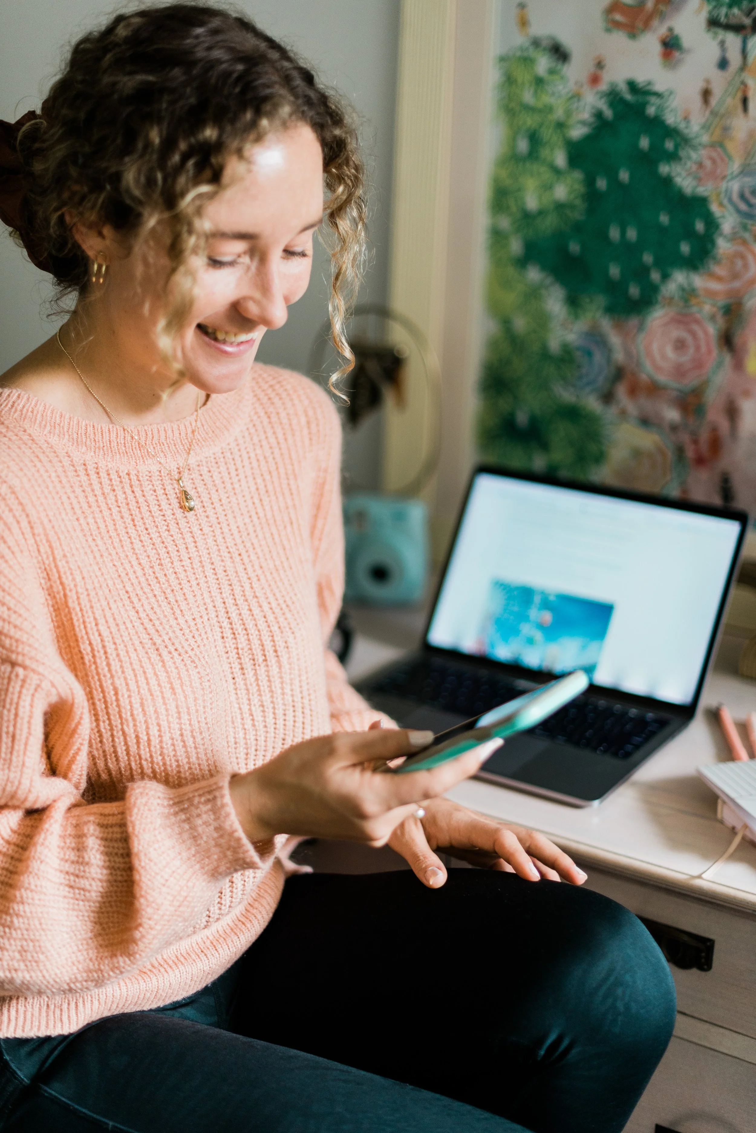 A young woman with curly hair, wearing a pink sweater, sitting and smiling while looking at her smartphone. There is a laptop, a notebook, and a pen on the desk beside her.