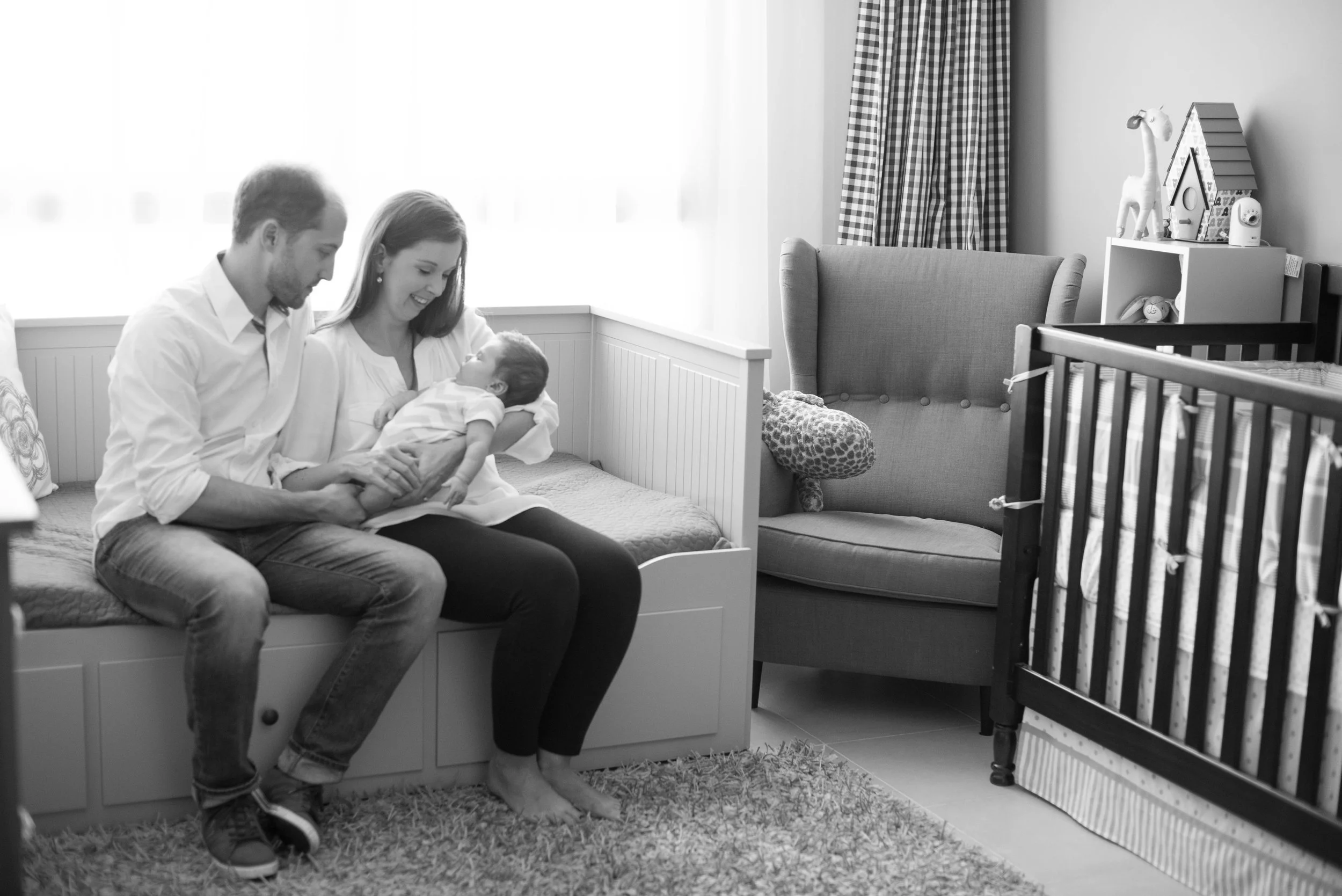 A family of three with a newborn sitting on a bed in a nursery, smiling and looking at the baby, with baby toys and stuffed animals in the background.