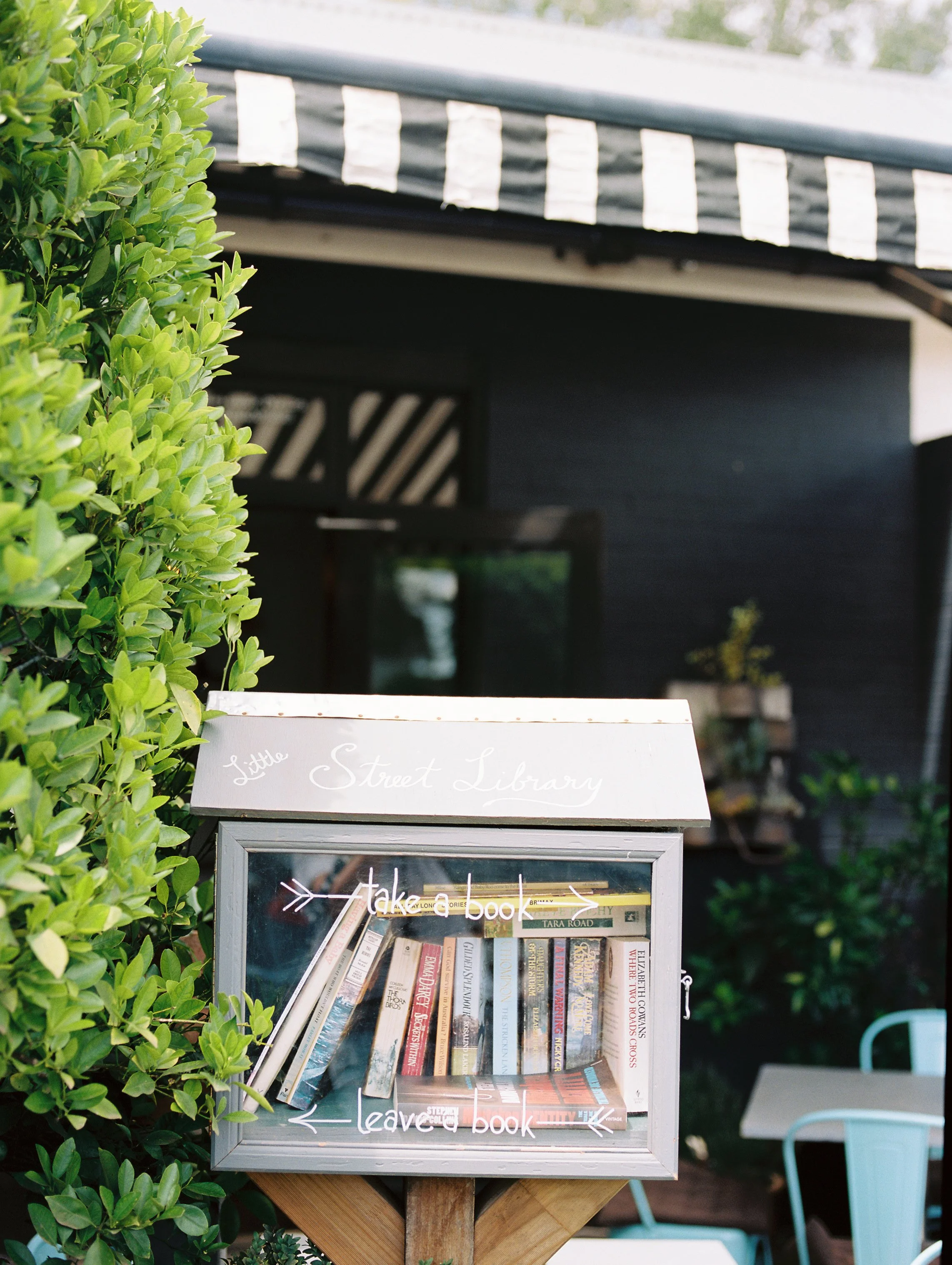 A small outdoor library box labeled 'Little Street Library' with a glass front, containing books, positioned next to a green bush. There are white metal chairs and a table in the background.