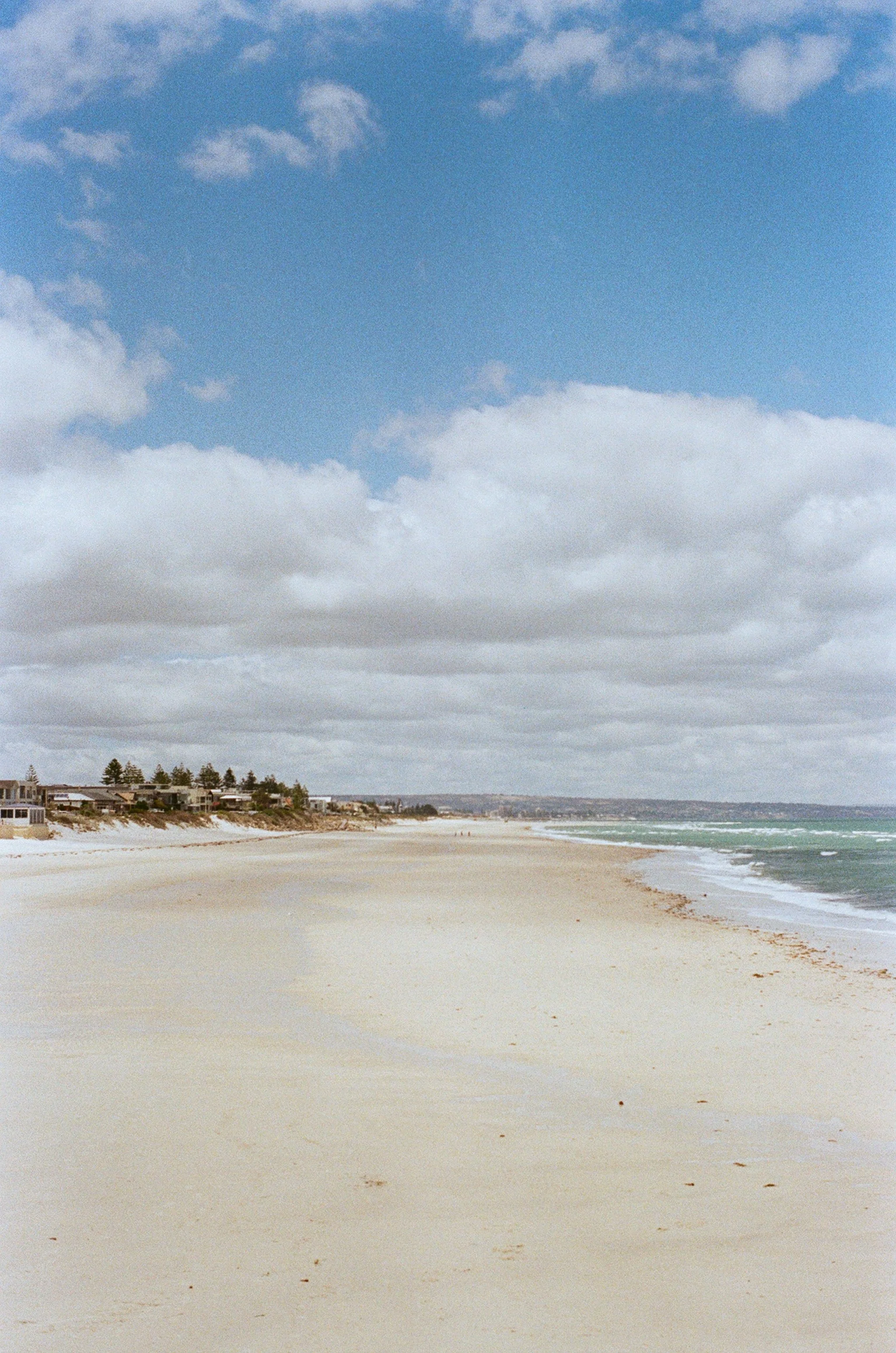 Empty sandy beach with houses in the distance and a cloudy sky over the ocean.