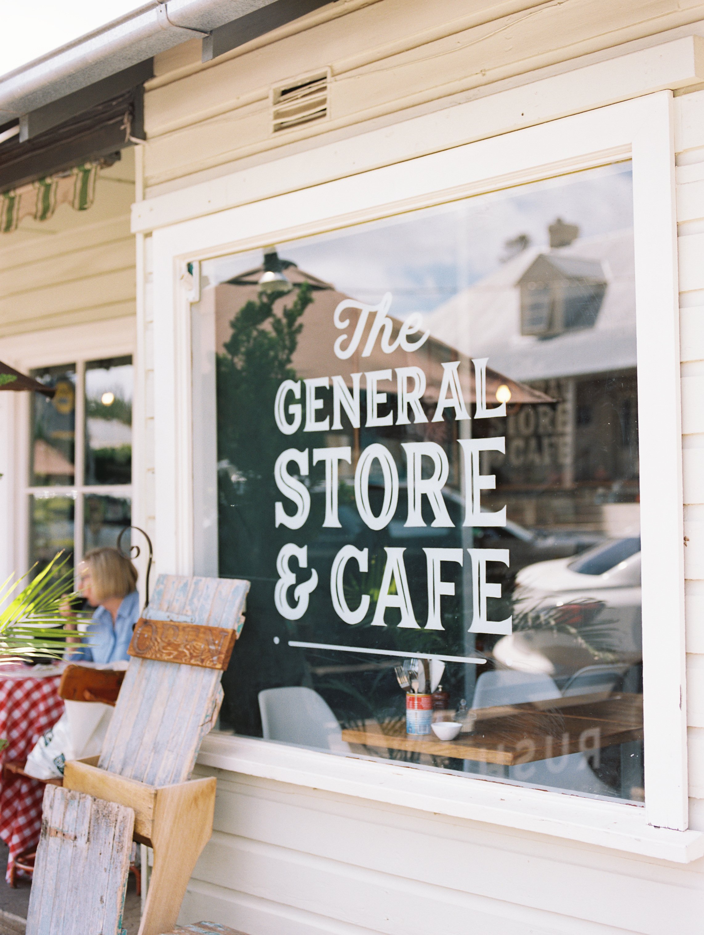 Exterior of a store and cafe with a large window displaying the sign 'The General Store & Cafe.' Reflection of buildings and cars in the window, with a person sitting outside at a table.