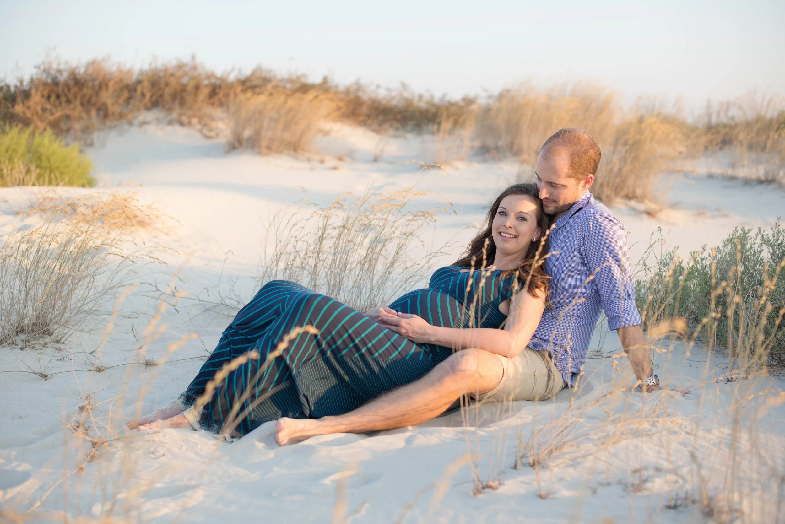 A couple sitting on the sand dunes, with the woman pregnant, smiling and embracing each other, surrounded by beach grasses at sunset.