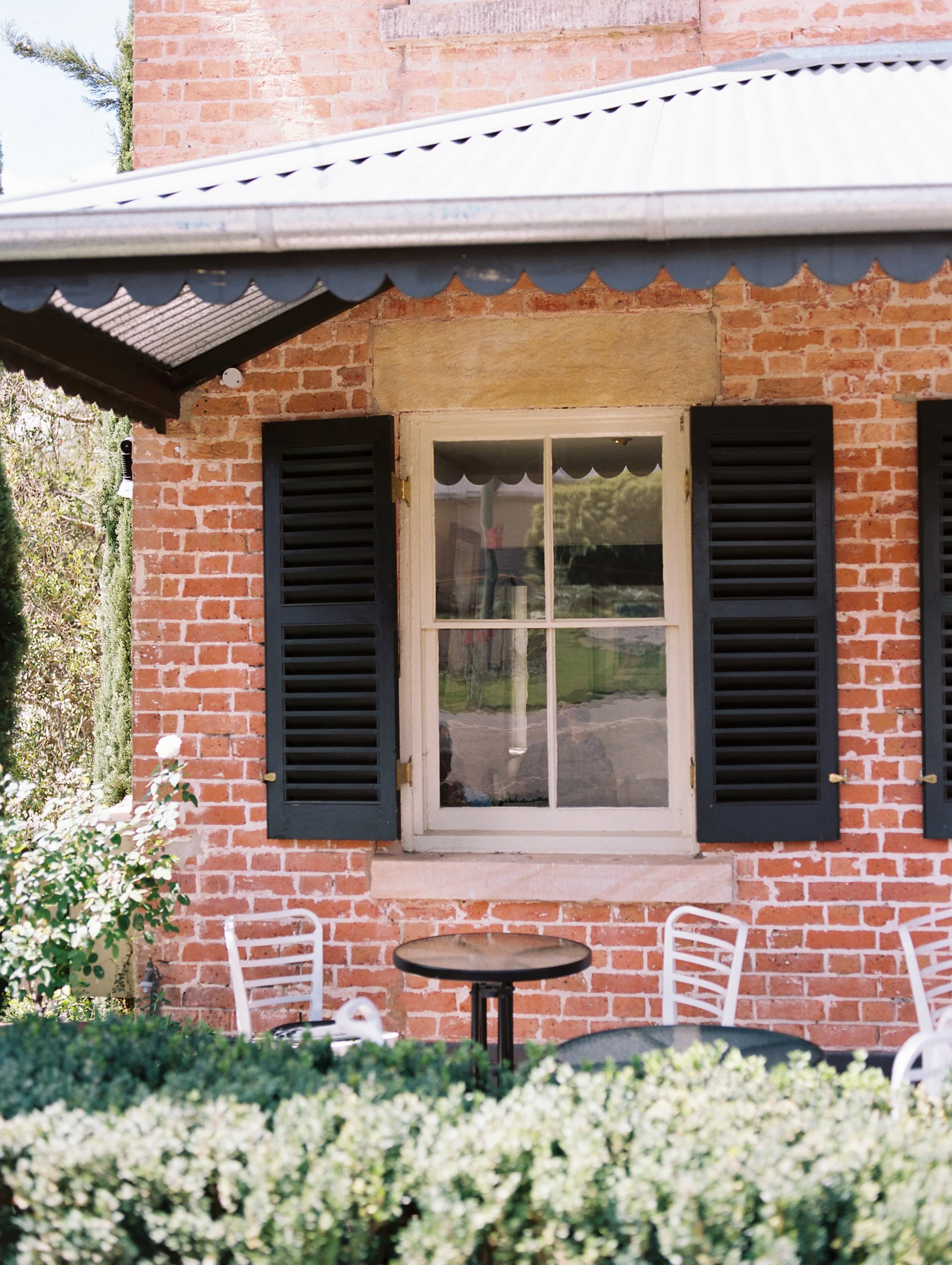A brick house exterior with a window flanked by black shutters, an awning overhead, and outdoor seating in front.