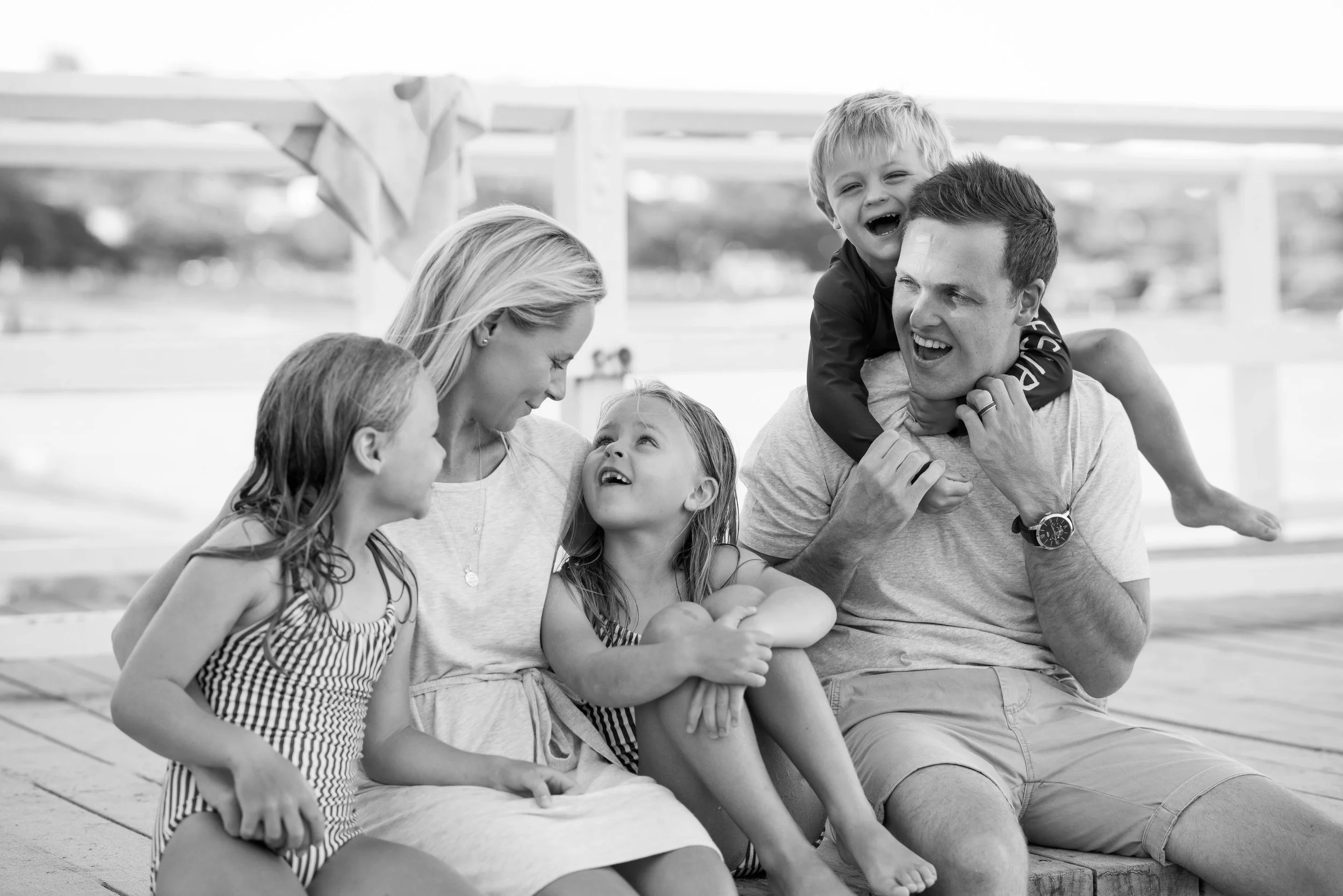 A family of six, including two adults and four children, sitting on a dock and smiling at each other. One child is on a man's shoulders, and a woman and three kids are engaged in conversation.