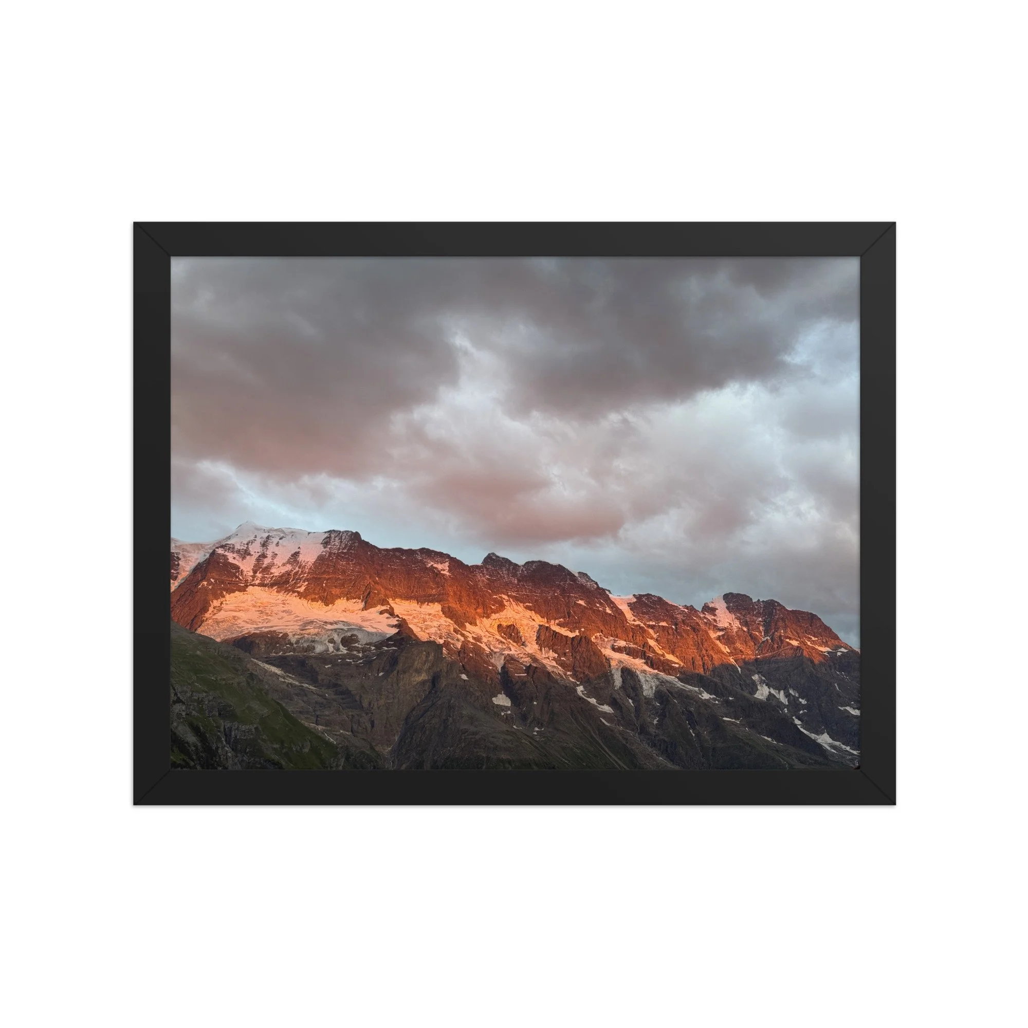 Warm beleuchtetes Bergmassiv mit Schnee unter grauen Wolken, Fotografie von Daniel Frei im schwarzen Rahmen. Warm light on snowy mountains beneath grey clouds, framed photograph.