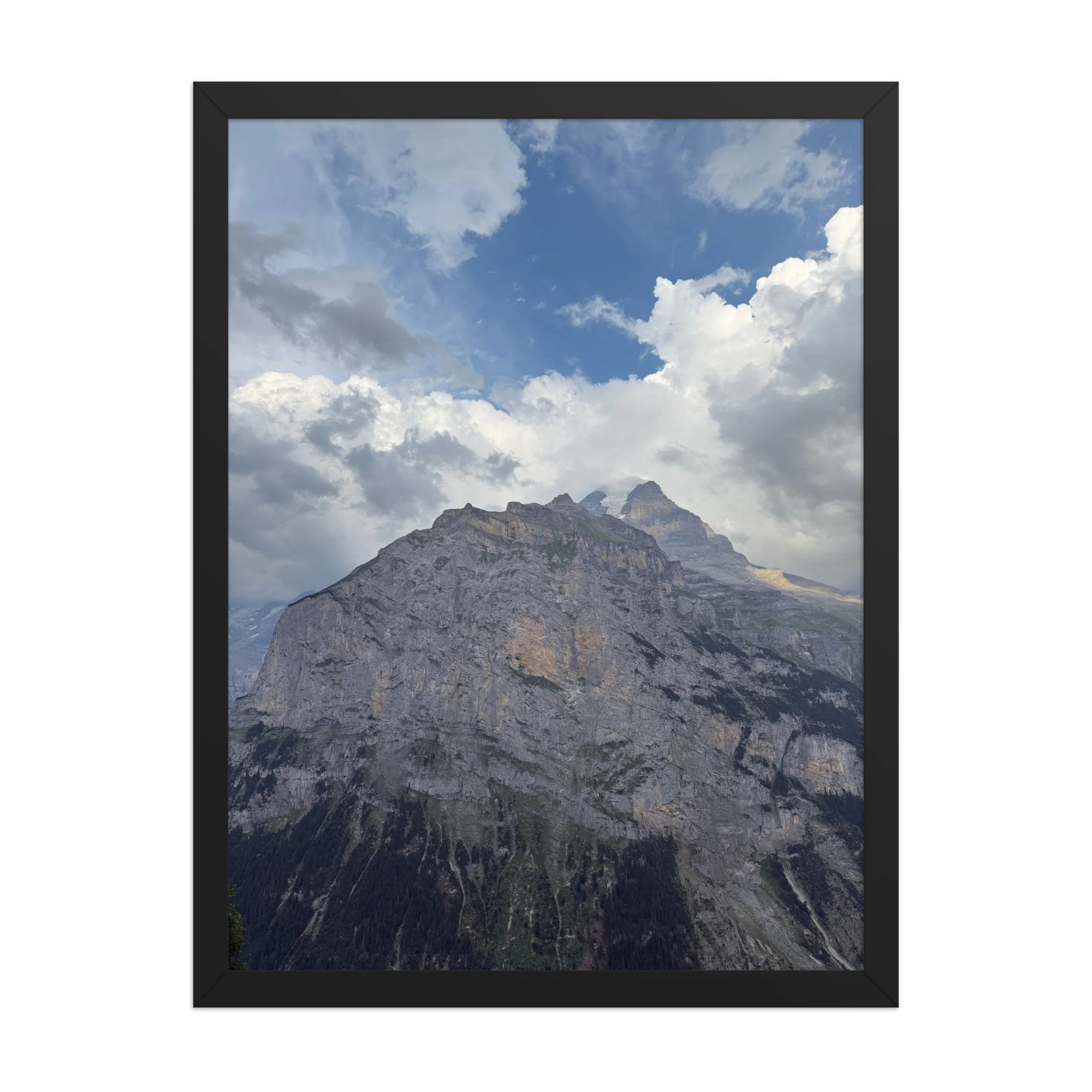 Felswand unter weissen Wolken und blauem Himmel, Fotografie von Daniel Frei im schwarzen Holzrahmen. Rock face beneath white clouds and blue sky, photograph in a black wooden frame.