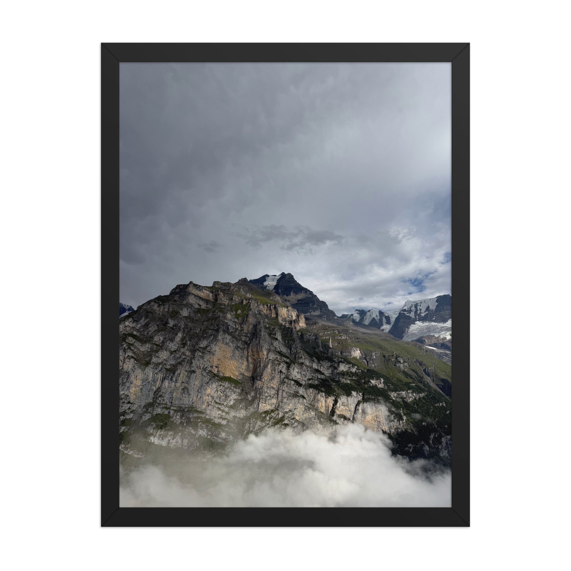 Felswände zwischen aufsteigenden Wolken und grauem Himmel, Fotografie von Daniel Frei im schwarzen Rahmen. Rock faces between rising clouds and a grey sky, framed photograph.