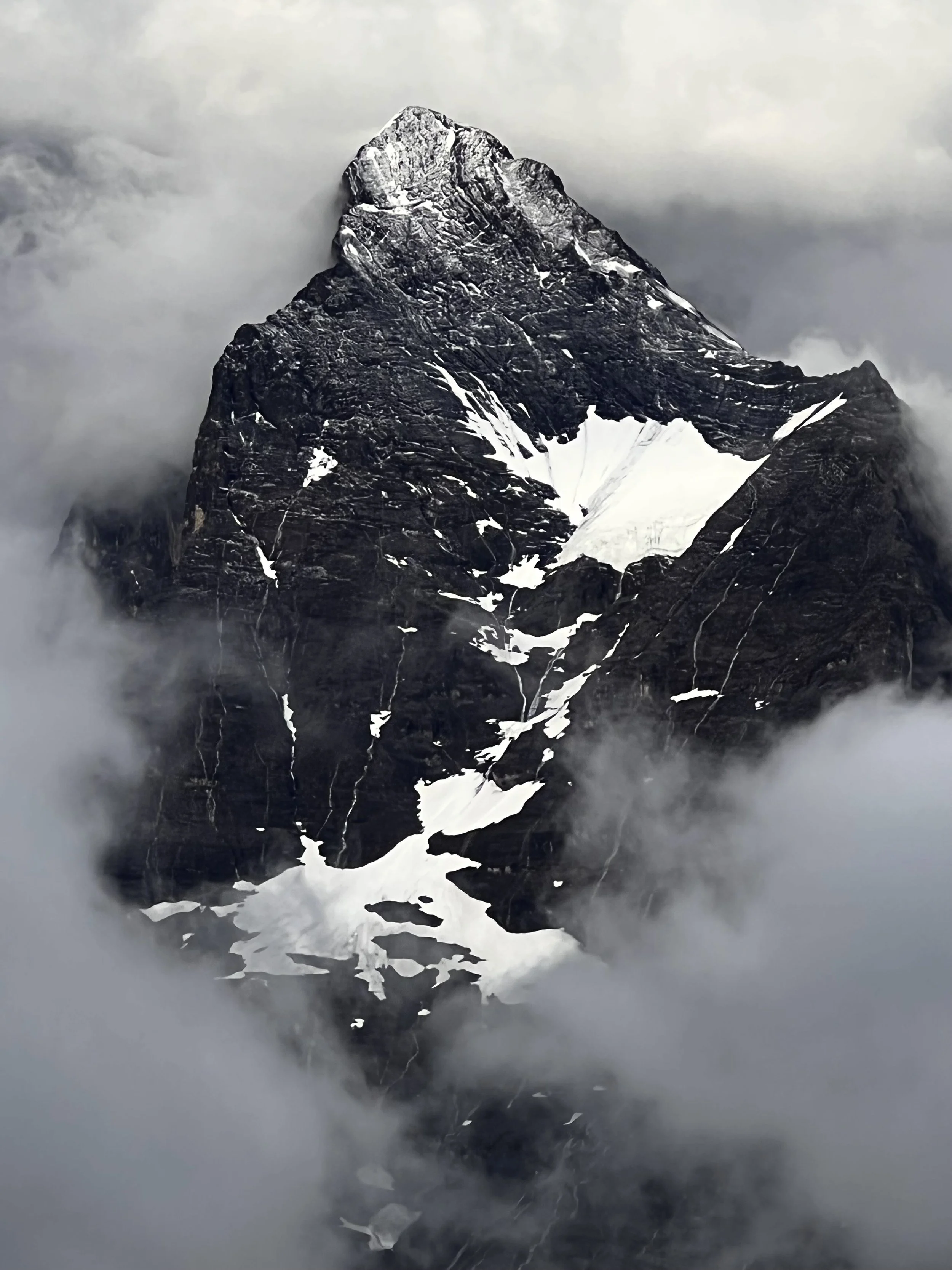 Dunkler Berggipfel zwischen Wolken und Schneefeldern.