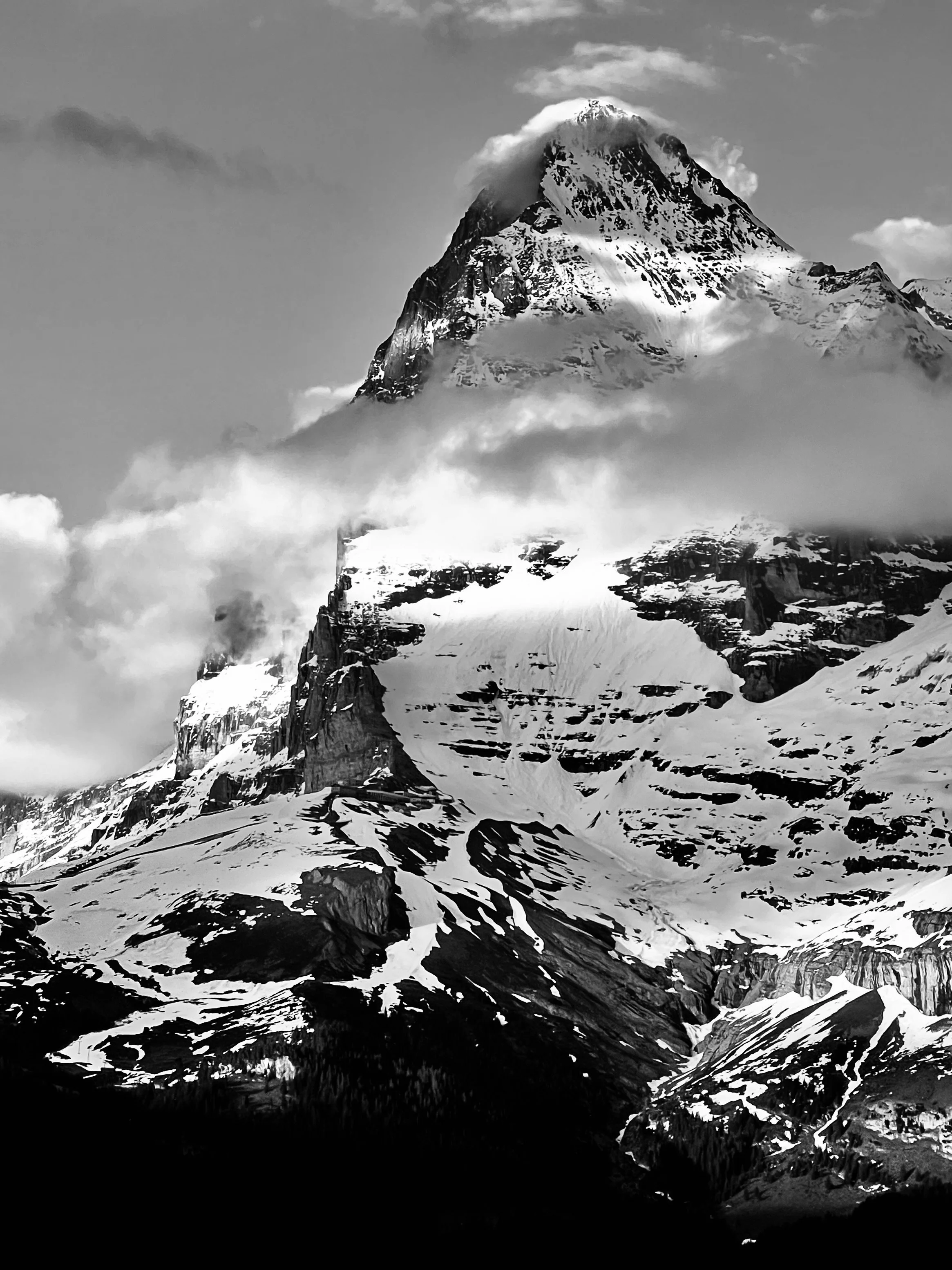 Wolkenbänder vor einer verschneiten Bergflanke in Schwarzweiss.