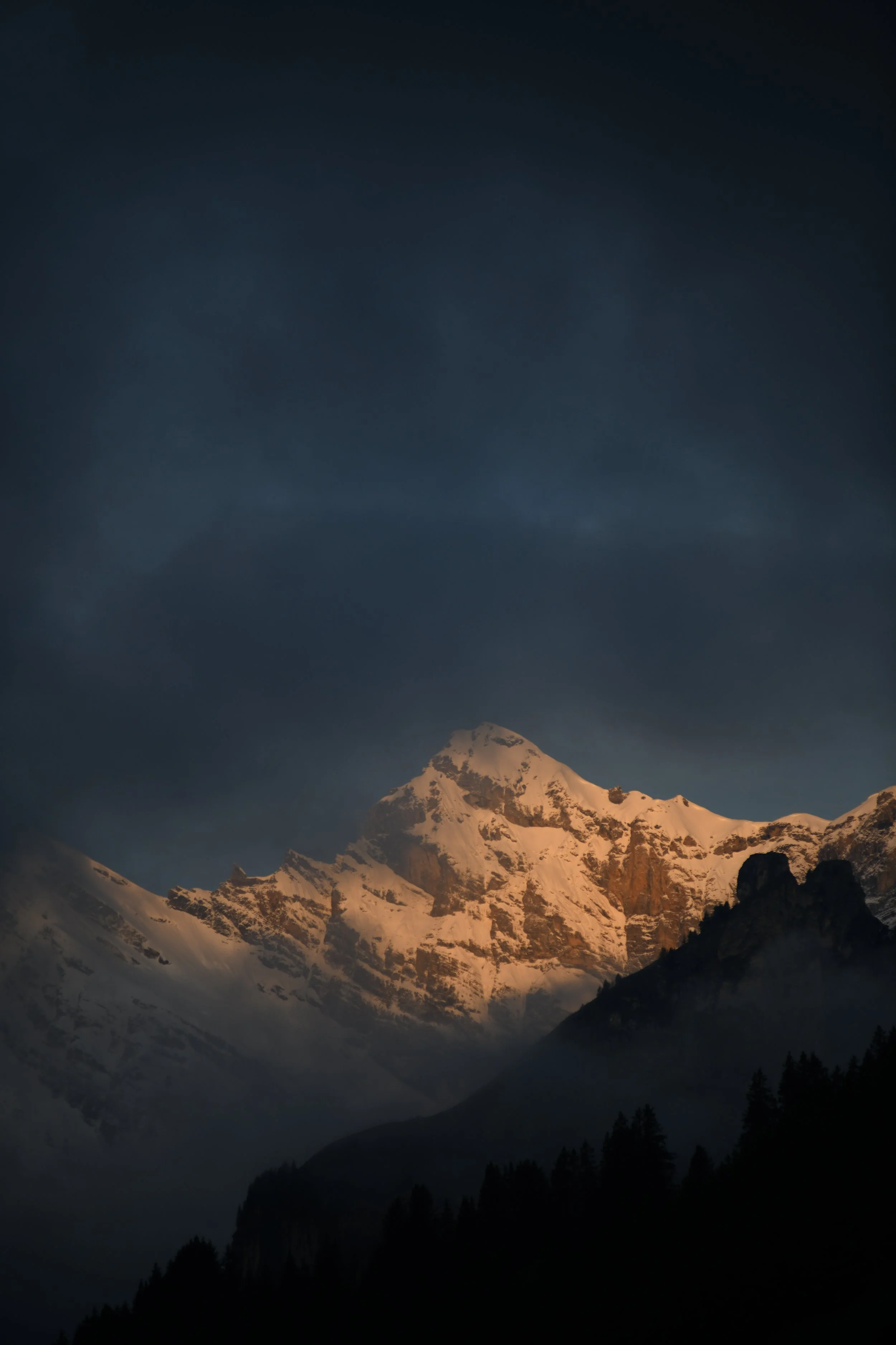 Lichtwechsel: Warmes Licht auf einer verschneiten Bergflanke unter dunklem Himmel. Fotografie von Daniel Frei.