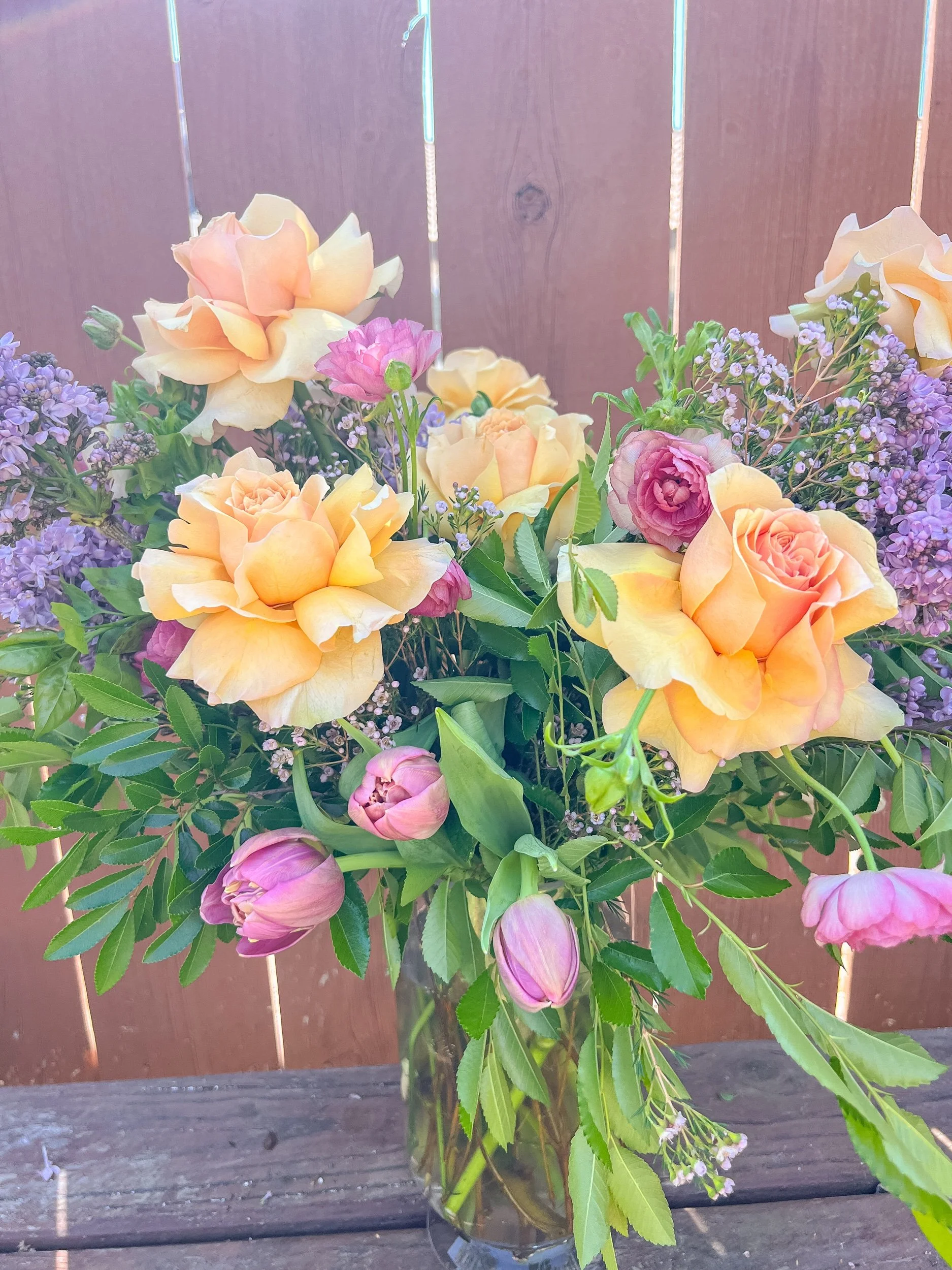 A bouquet of pastel-colored roses and purple flowers in a glass vase on a wooden table.