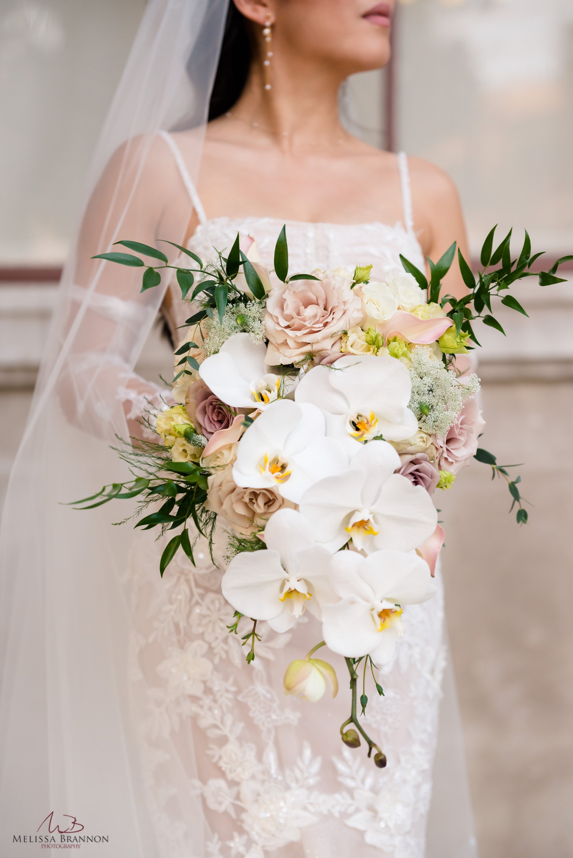 Bride in lace gown holding white and pink bouquet with orchids and greenery.