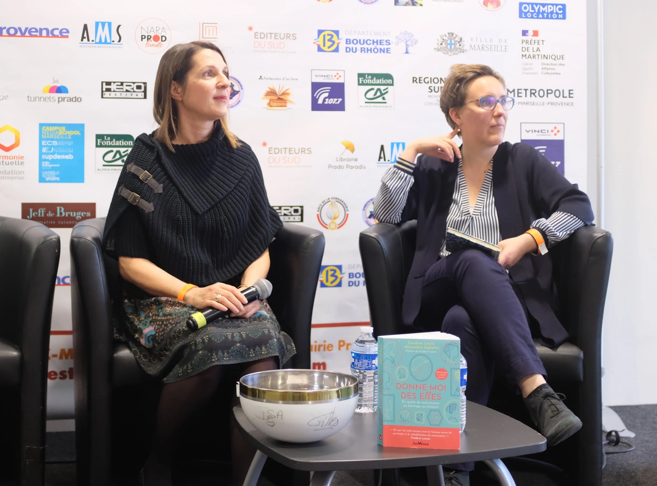 Deux femmes assises sur des fauteuils noirs avec un fond de logos, l'une tenant un microphone. Une table avec deux bouteilles d'eau et un livre intitulé "Donne-moi des elles".