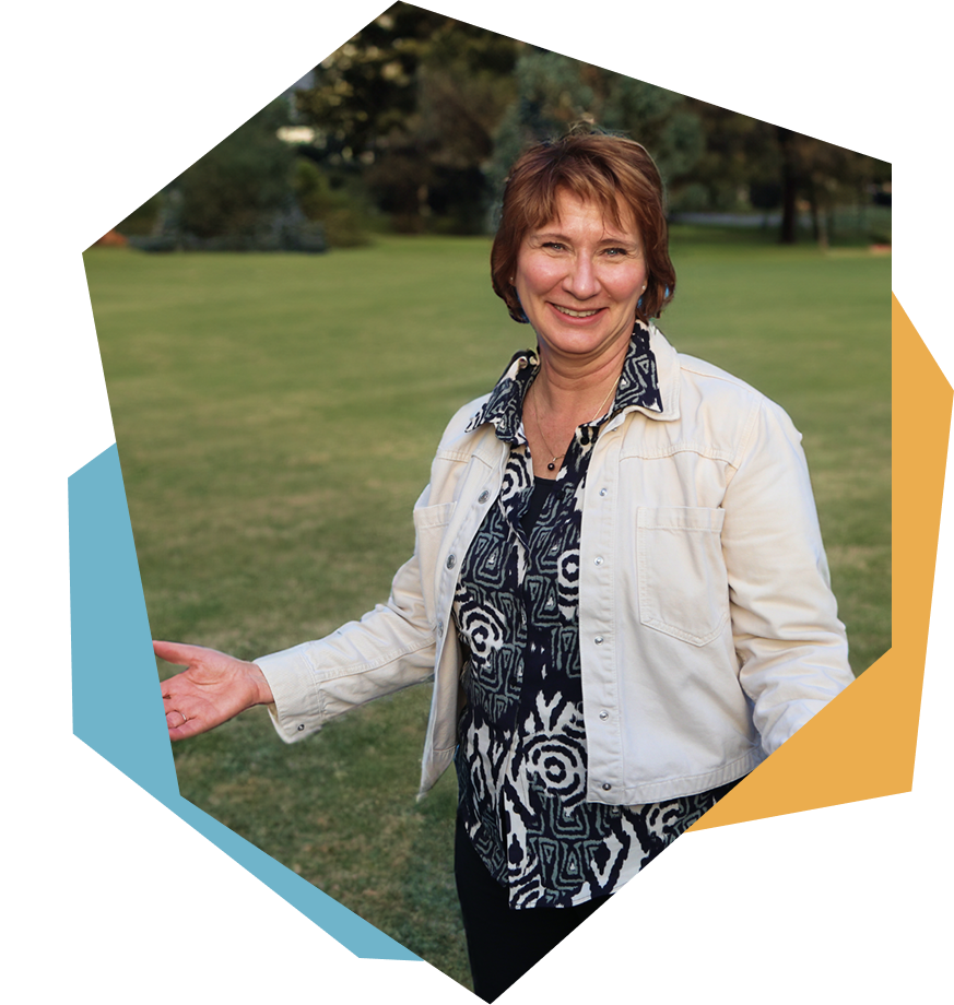 Vrouw staat in een park, glimlacht, draagt een witte jas en een zwart-wit gebladerd top, met groene gras en bomen op de achtergrond.