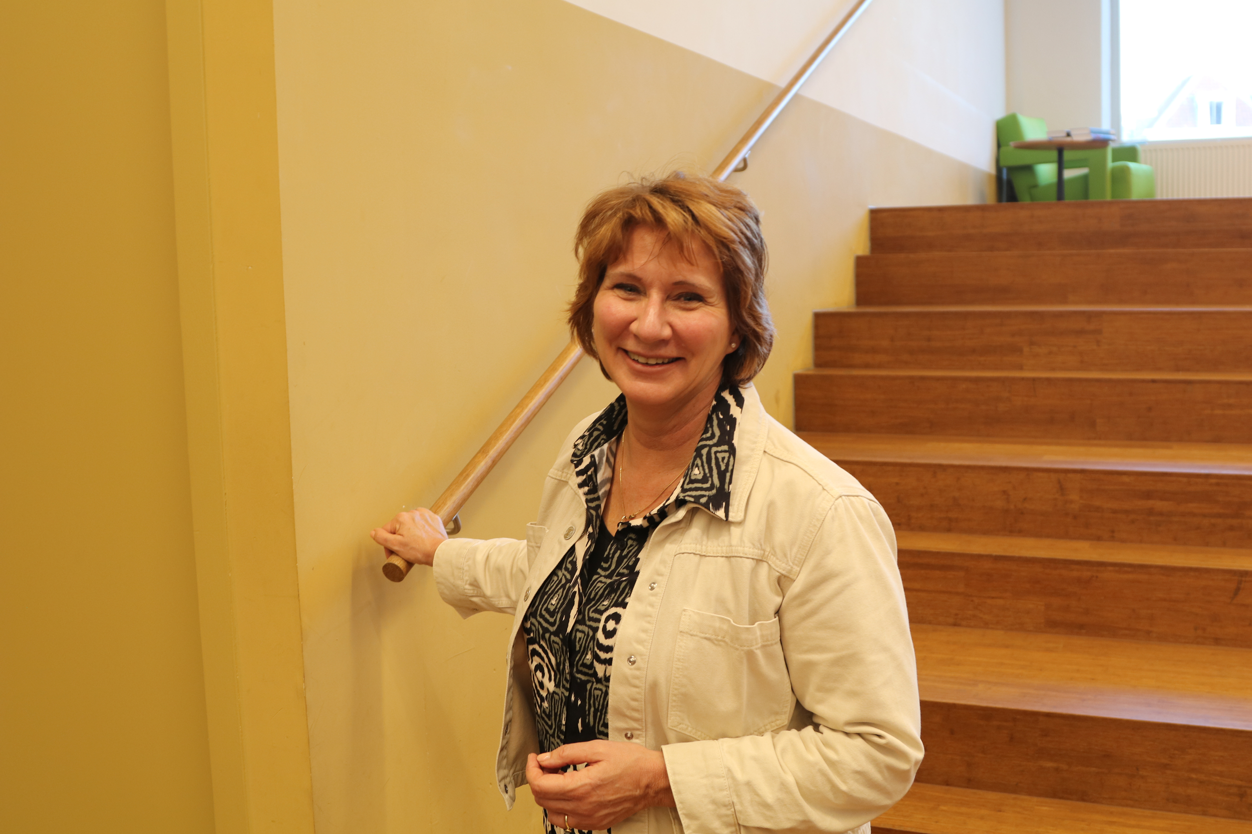 Monique hoogduin die glimlacht terwijl ze zich vasthoudt aan een muur en een trap opgaat, in een interieur met gele muren en houten trappen.